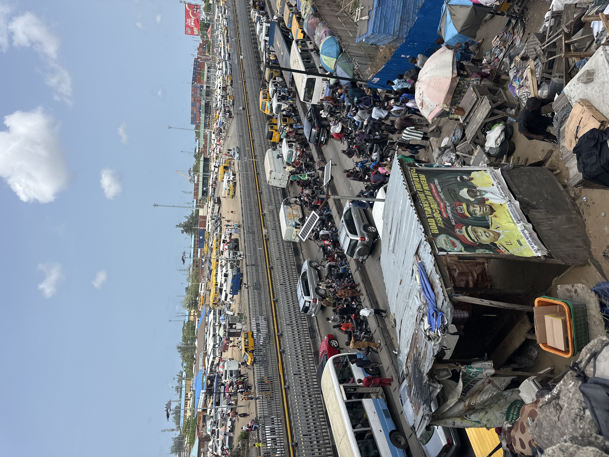 The image depicts a congested street scene in Lagos, Nigeria. The road is densely packed with yellow taxis, buses, and other vehicles, indicating heavy traffic. Numerous people are walking along the sides of the road and among the vehicles. The scene appears to be taken from an elevated position, possibly overlooking the street.
To the right of the road is an area with makeshift structures, perhaps shops or dwellings constructed with corrugated metal and other materials. A poster on one of the structures mentions the "Road Transport Employee's Association of Nigeria," suggesting a connection to transport workers. The poster also mentions "Mile 2 Promise Phase 1 Chapel, Amuwo Zone," thus identifying the location in the city. The environment seems to be a mix of commercial activity and residential areas. The sky above shows a bright, sunny day.