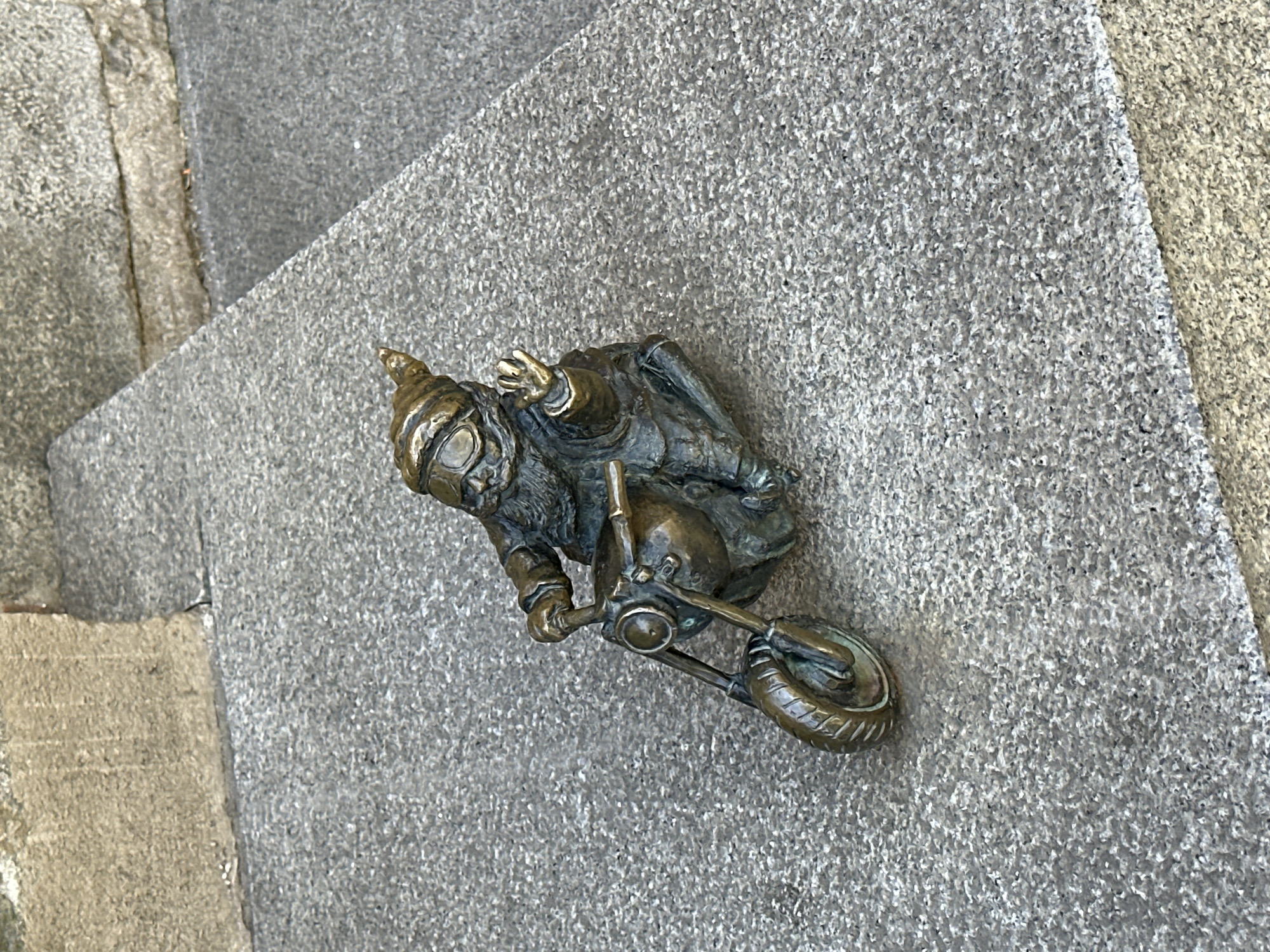 A bronze sculpture of a gnome wearing goggles and a pointed hat, perched on a motorcycle, is affixed to a gray, textured stone surface. The gnome has one hand raised in a waving gesture. The location is the city of Wrocław, Poland, where numerous such gnome sculptures are placed around the city. The stone surface appears to be part of a building facade or paving.