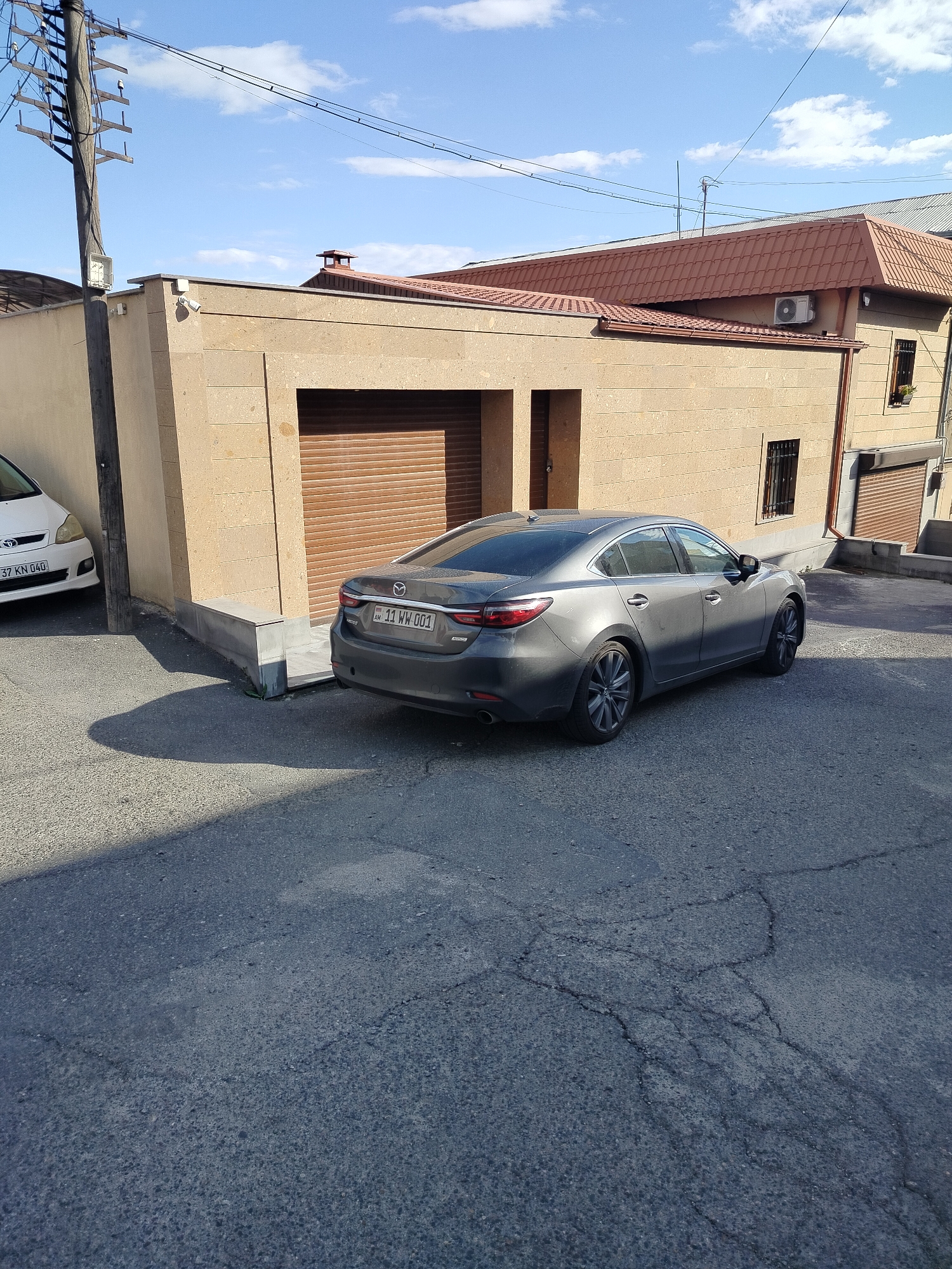 The image shows a gray Mazda 6 sedan parked on a paved street in Yerevan, Armenia. The car's license plate is "11 WW 001". The car is parked next to a light brown stone building with a closed garage door. A white Toyota is parked on the other side of the building. Overhead power lines and a utility pole are visible in the background. The sky is blue with scattered clouds. Another building with a brown roof is in the distance.