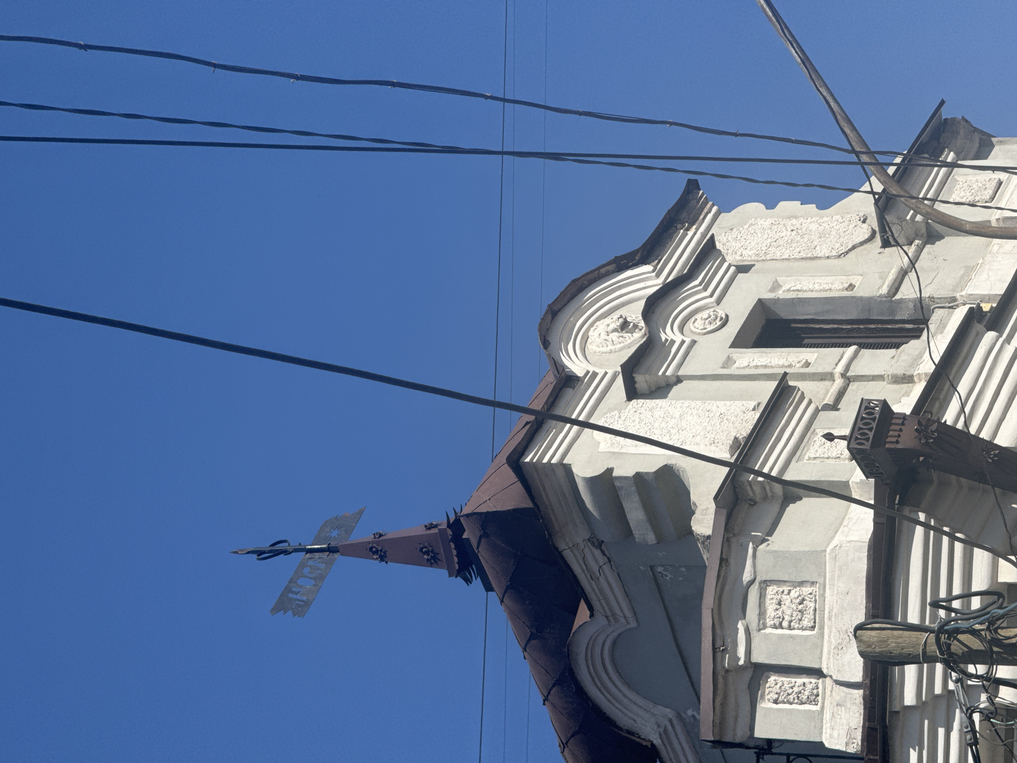 The image presents an upward view of an ornate building facade against a bright, clear blue sky, interwoven with numerous dark power lines. The building, possibly an older structure, features a light-colored stucco or stone facade with intricate decorative elements, including curved mouldings, raised textured panels, and recessed sections. Its roofline is characterized by dark, possibly metal, tiles or cladding, culminating in a distinctive peak.
Several thick, dark power lines stretch across the upper portion of the frame, crossing horizontally and diagonally. On the left side of the building's peak, a decorative metal finial or weather vane is visible. It is dark-colored, with an arrow-like shape and a prominent horizontal crossbar. Etched or cut into this crossbar is text that appears to read "NOCHE," accompanied by a starburst symbol.
The scene is illuminated by bright daylight, suggesting a sunny day with no clouds. No people or specific activities are depicted. On the far right, part of a utility pole or similar wooden support structure is visible, with a bundle of wires attached to it. The architectural style hints at a historical urban setting, consistent with its location in Krasnoyarsk, Russia.