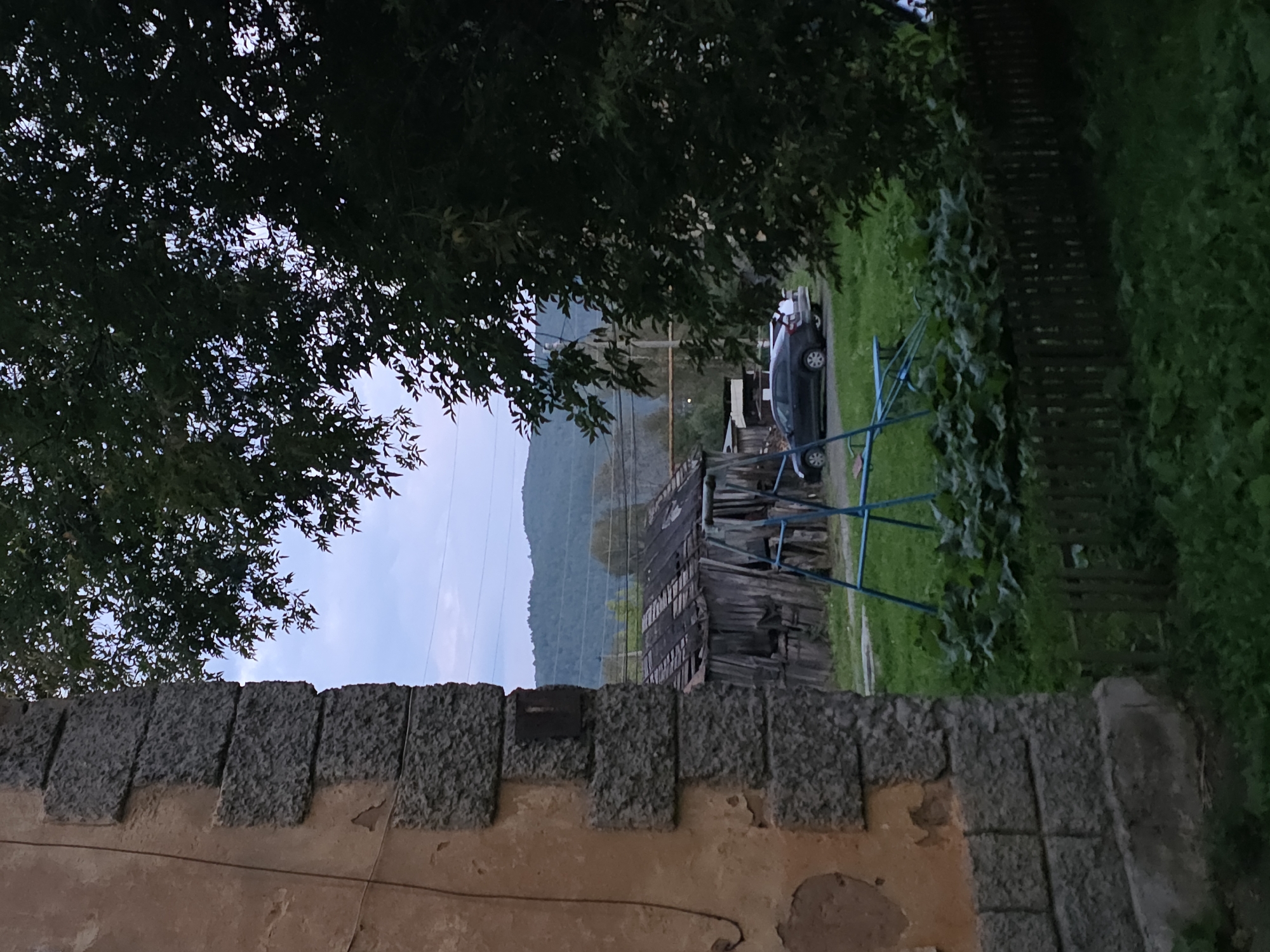 This image depicts a rural or semi-rural scene, likely on the outskirts of Magnitka, Russia, given the context. In the foreground to the left, a weathered stone pillar of a building is visible, showing signs of age with chipped paint and textured stone blocks. To the right, dense green foliage from a large tree partially obscures the view.
Beyond these elements, a dilapidated wooden structure with a damaged roof is present. In front of this structure, a blue metal playground swing set is set up on a grassy area. A dark-colored sedan is parked on a dirt or gravel surface behind the swing set, with another vehicle partially visible behind it. In the background, rolling hills covered in trees are seen under a pale, overcast sky, with power lines stretching across the horizon. The overall impression is one of a somewhat neglected rural setting, possibly a backyard or a communal area. There are no people visible in the image.