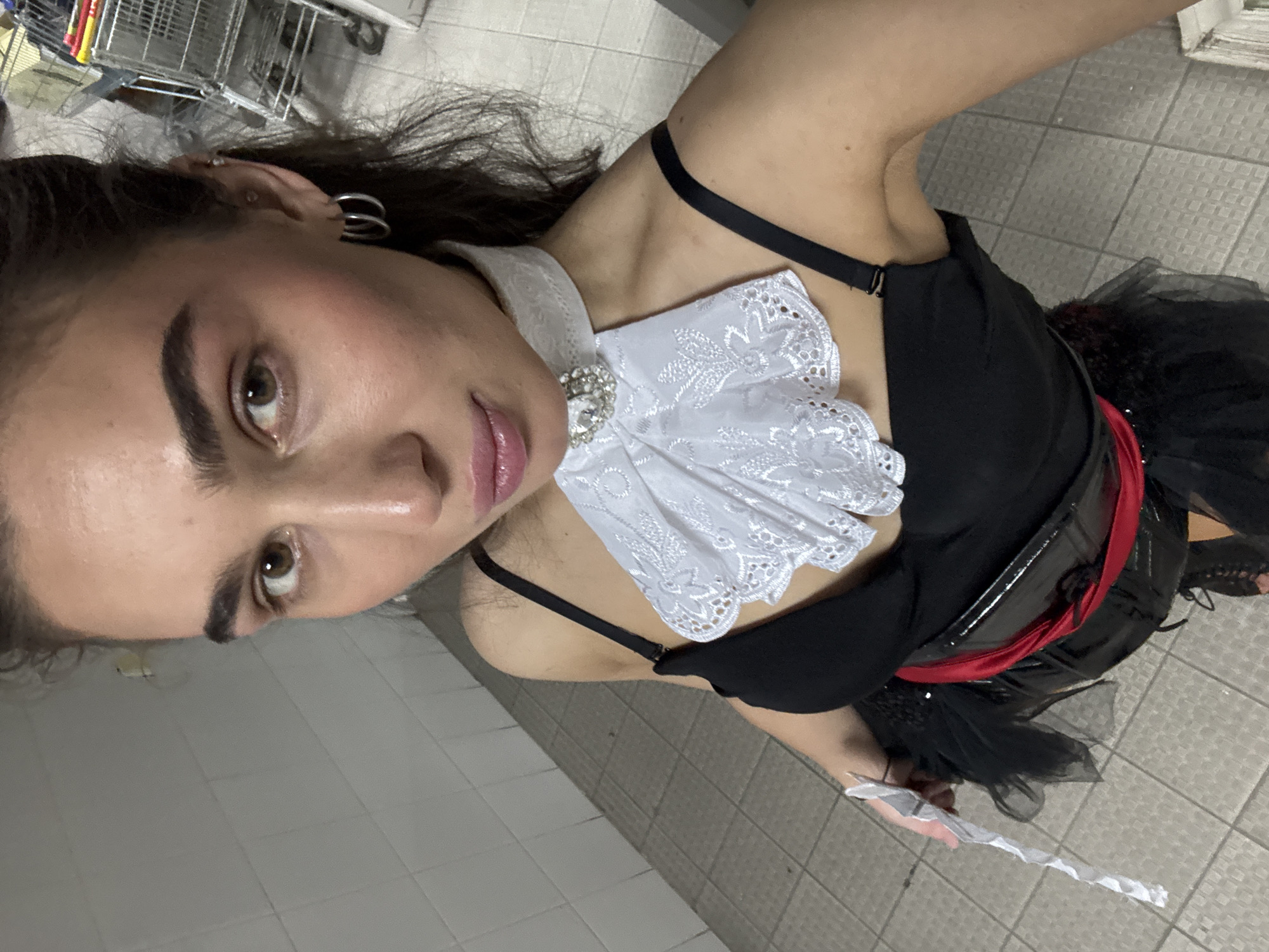 The image shows a woman in a costume, possibly for a performance or event, taking a selfie. She has dark hair, brown eyes, and is wearing a white lace collar with a jeweled embellishment, a black top with thin straps, and a black tutu-like skirt with a red sash. She is holding a thin, white object in her left hand. The background appears to be a tiled wall and floor, suggestive of a public restroom or backstage area. In the far background, a shopping cart is visible. The scene has the appearance of being indoors.
