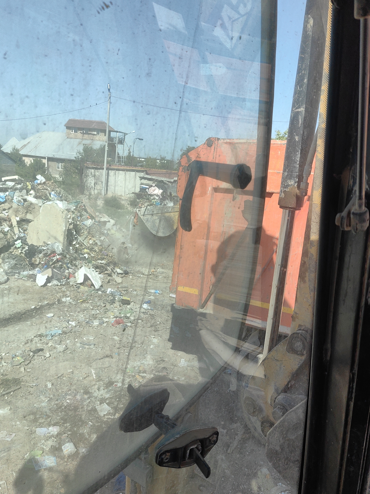 The image, taken from inside a vehicle cabin, depicts a landfill in Shymkent, Kazakhstan. A large orange garbage truck is positioned in the foreground, with its collection mechanism extended and actively scooping debris from a large pile of trash. The ground is covered in a mix of dirt, debris, and scattered refuse, including plastic bottles, paper, and larger discarded items. In the background, a residential building with a pitched roof and several windows is visible, along with trees and what appears to be a concrete structure. The sky is clear and blue, suggesting daytime. The scene conveys a sense of industrial waste management and the challenging environmental conditions often associated with such sites.