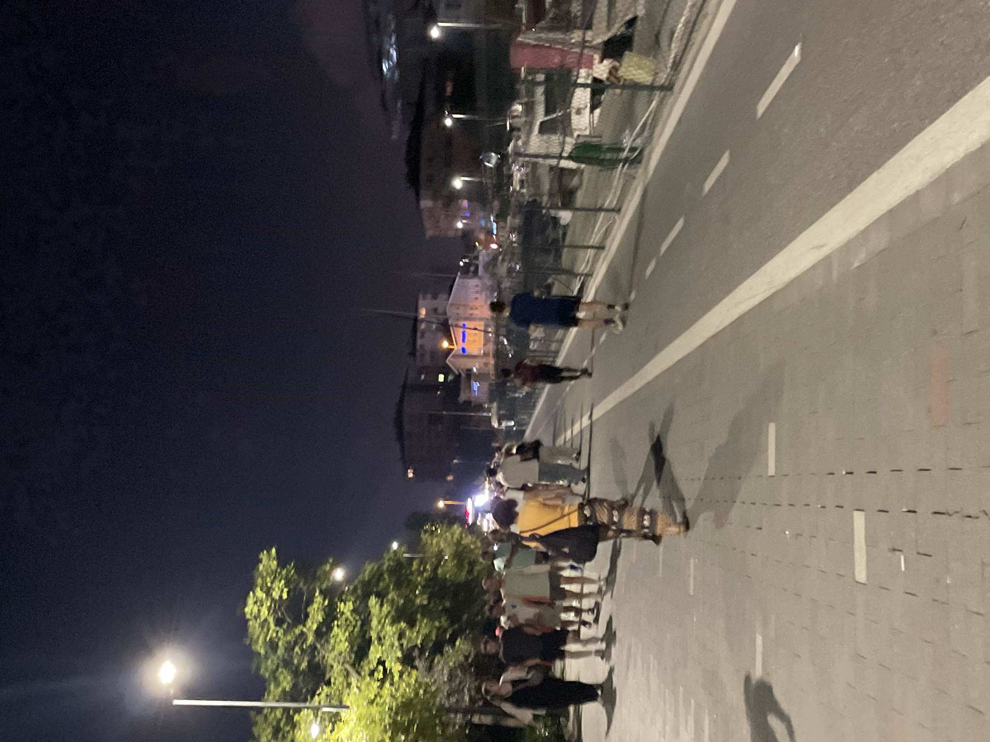 In Kadıköy, Turkey, at night, a group of people stroll along a paved walkway adjacent to a road. The walkway is illuminated by streetlights and the ambient light from buildings in the background. Several individuals, dressed in casual attire, are walking in the same direction. To the right of the walkway, there is a road with painted lane markings, and beyond that, a fence with some debris. In the distance, buildings and what appear to be boats are visible. The overall scene depicts a common urban nighttime activity.