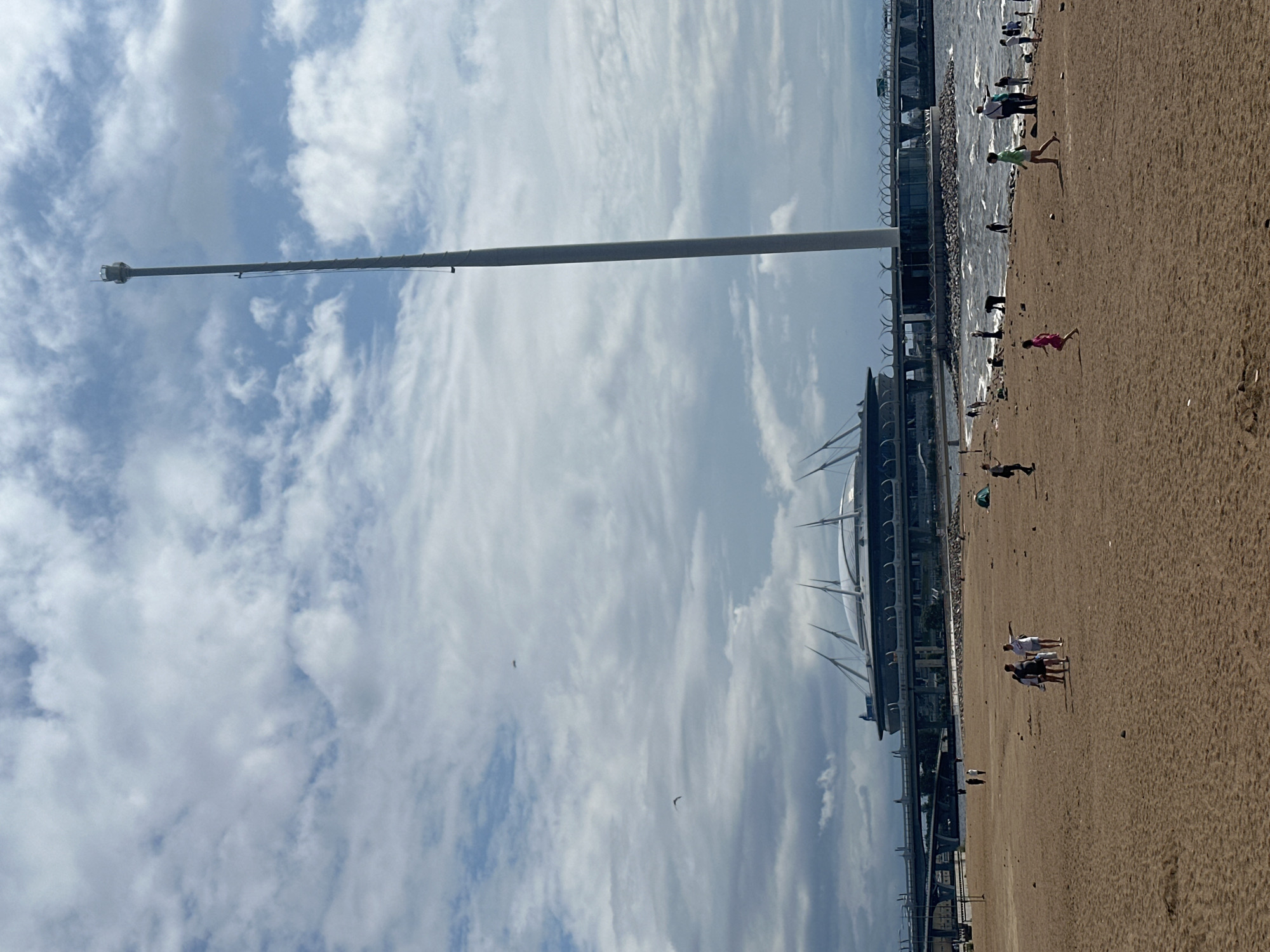 An outdoor, eye-level, wide shot captures a beach scene with a stadium and a tall tower in the background under a cloudy sky. The foreground is a sandy beach with a group of people scattered across it. In the middle ground, the choppy waters of a body of water are visible, with more people along the shoreline and some further out. The background features a large, modern stadium with a distinctive roof structure and a very tall, slender tower. A bridge or elevated roadway also spans across the scene behind the stadium. The overall atmosphere is casual, with people enjoying the beach on a cloudy day.