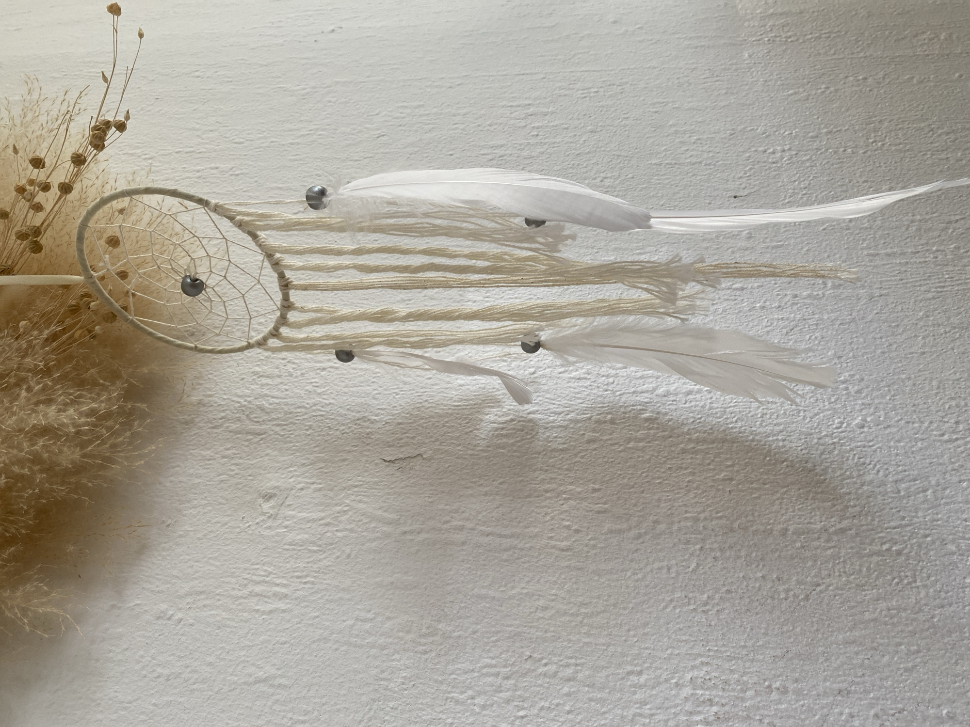 The image depicts a dreamcatcher hanging on a textured, off-white wall. The dreamcatcher has a circular frame woven with white string, and several strands of the same string hang down from it. White feathers are attached to some of these strands, and small, silver-colored beads are interspersed throughout. Above the dreamcatcher, a cluster of dried, feathery plants is visible. The wall itself appears to be painted or stuccoed, with a rough, uneven surface. The lighting suggests an indoor setting, possibly a home or a shop. The image does not contain any information that would allow for localization in Bad Sachsa, Germany.