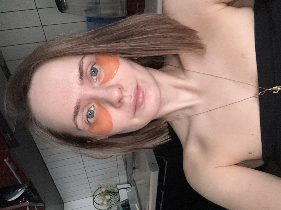A woman wearing orange under-eye patches poses for a selfie. She has light skin, blue eyes, and short, brown hair. A delicate gold necklace hangs around her neck. She is wearing a black top. The background includes a tiled wall and other kitchen elements. The photograph is taken from a slightly high angle and rotated counterclockwise.