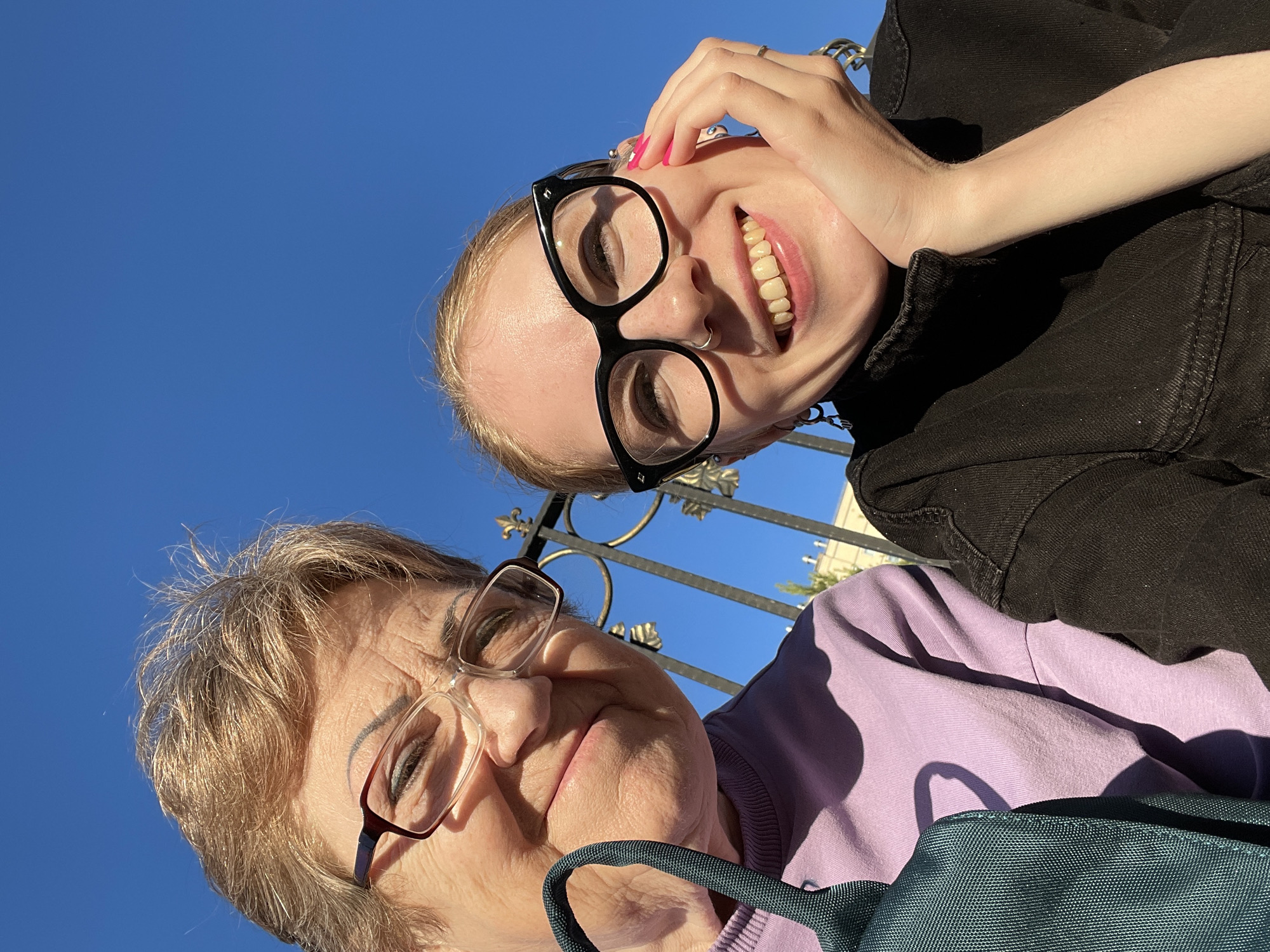 Two individuals, a younger person and an older woman, are posing for a selfie against a bright blue sky. The younger person is wearing black-rimmed glasses and a black jacket, with a visible nose piercing and pink nail polish. They are holding their hand near their face, smiling broadly. The older woman is wearing glasses and a light purple sweater. A blue tote bag is also visible. Behind them is a wrought iron fence. The location is Izhevsk, Russia.
