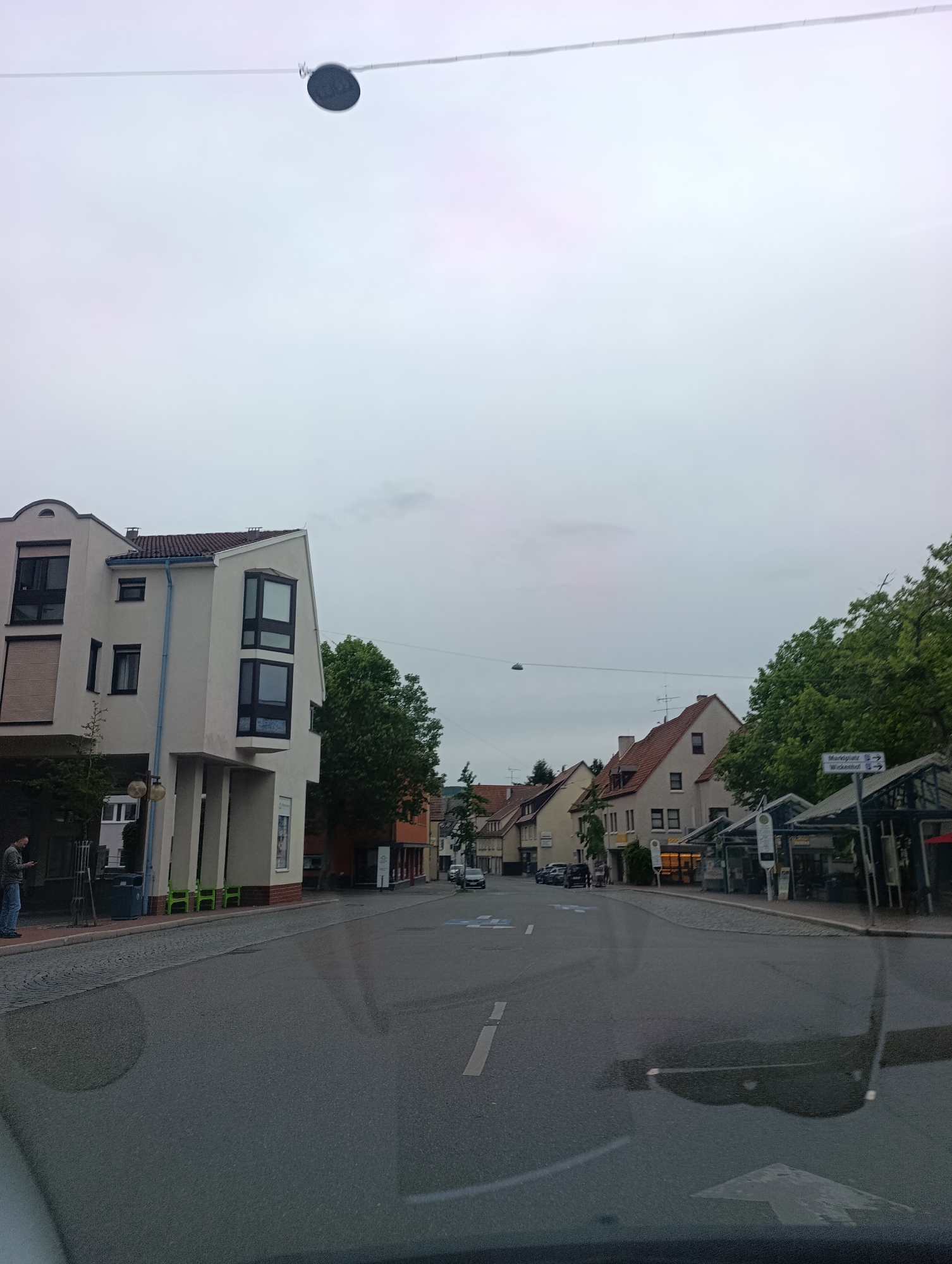 The image shows a street scene in Pfullingen, Germany, on a cloudy day. On the left side of the street is a modern, multi-story building with a white facade and dark windows. A man in a grey jacket and blue jeans is standing on the sidewalk in front of the building, looking at his phone. To the right of this building, the street continues, lined with traditional German houses with red-tiled roofs. Several of these houses have shops on the ground floor. In the foreground, the street is wet, possibly from recent rain, and a reflection of the sky and surrounding buildings can be seen on the asphalt. There are some parked cars on the street. Signs indicate directions to "Marktplatz" and "Wickenhof." Trees with green foliage are visible throughout the scene. A round, dark object is suspended from a wire in the upper part of the frame.