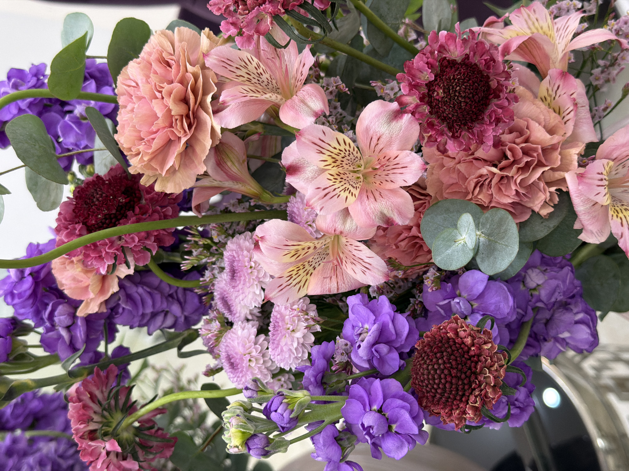 A close-up photograph depicts a bouquet of assorted flowers. The arrangement includes peach-colored carnations, pink alstroemeria with speckled markings, purple stock flowers, and dark red scabiosa. Green eucalyptus leaves are interspersed within the arrangement. The photograph focuses on the texture and color variations of the flowers. There are no indications or clues in the photograph about the location of Moscow, Russia.