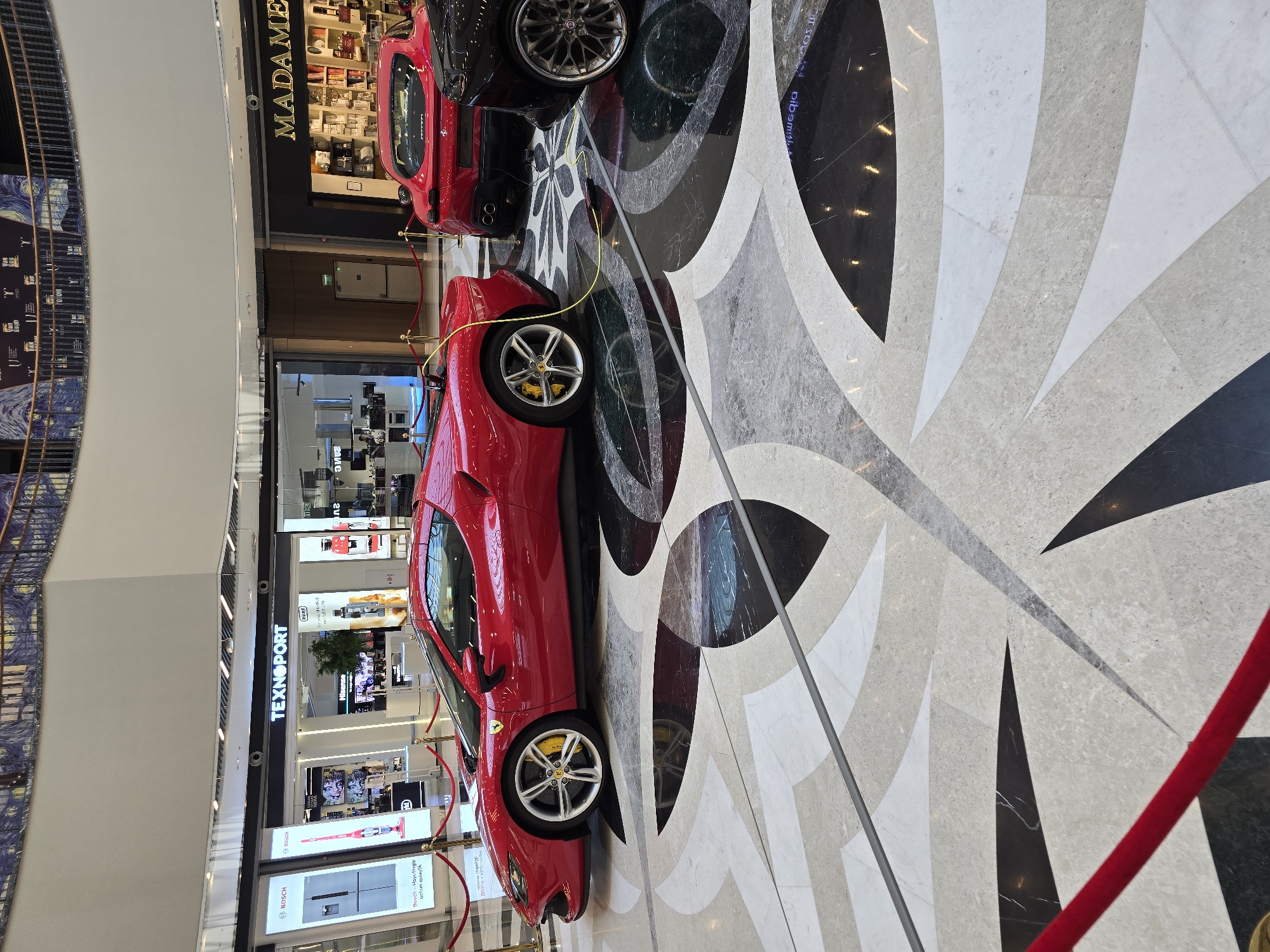 The image depicts a showroom display of red Ferrari sports cars on a patterned marble floor in a shopping mall. In the background, storefronts for "TEXNOPORT" and "MADAME" are visible. The overall environment suggests an indoor commercial space, likely a luxury retail setting. The scene is static, with no people actively engaged in the image. The cars are the central focus, presented as exhibits.