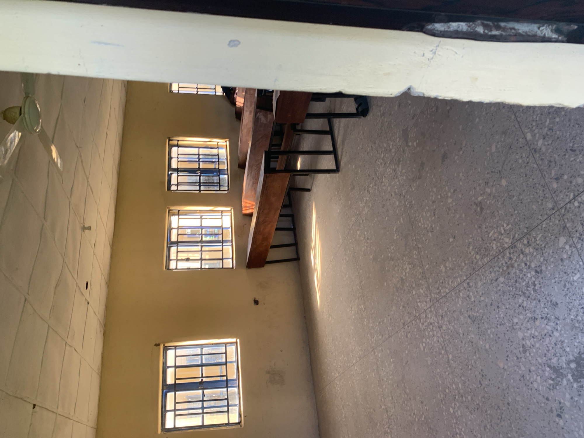 This image depicts the interior of a classroom located in Jalingo, Nigeria. The room is sparsely furnished, with rows of wooden desks and benches, some of which appear to be older and weathered. Sunlight streams through barred windows, casting long shadows across the speckled grey floor. A ceiling fan is visible, suggesting the need for ventilation in the climate. The walls are painted a pale yellow and show some signs of wear and tear. The overall impression is one of a functional, perhaps older, educational space. There are no people visibly present or interacting in the scene.