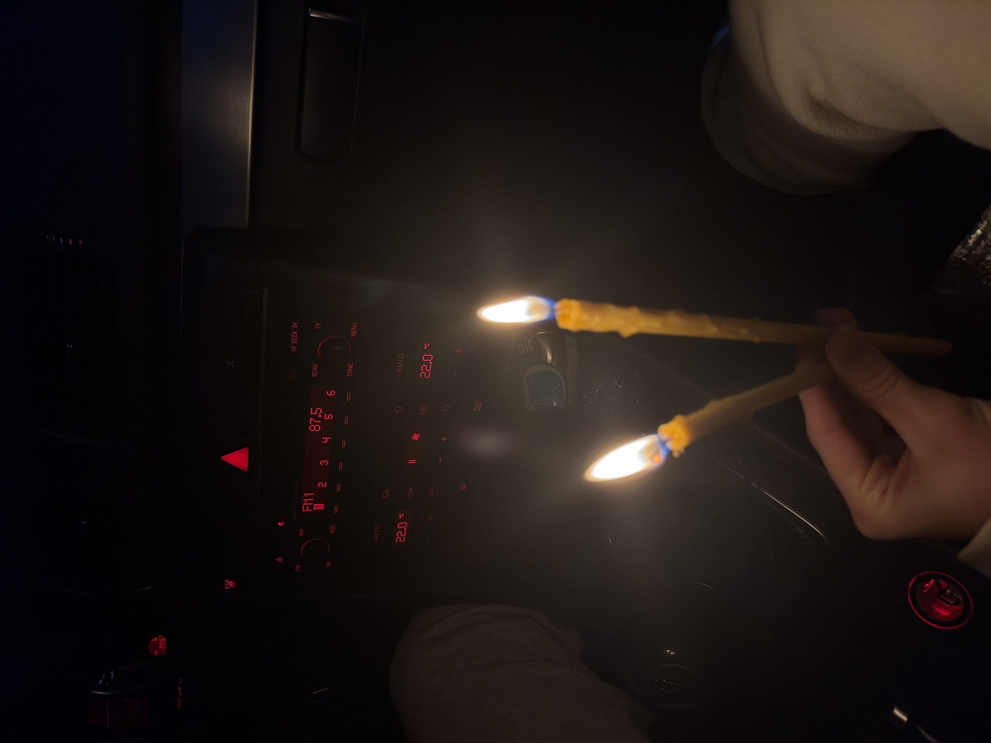 Here is a forensic description of the image:
The image is taken from inside a vehicle at night. The dashboard is visible, with red illuminated buttons and displays. The radio is tuned to FM1 at 87.5. The climate control is set to AUTO and 22.0 degrees. Two lit candles, likely ear candles, are being held in someone's hand. The location of Milevo, Bulgaria, is not discernible from this image.