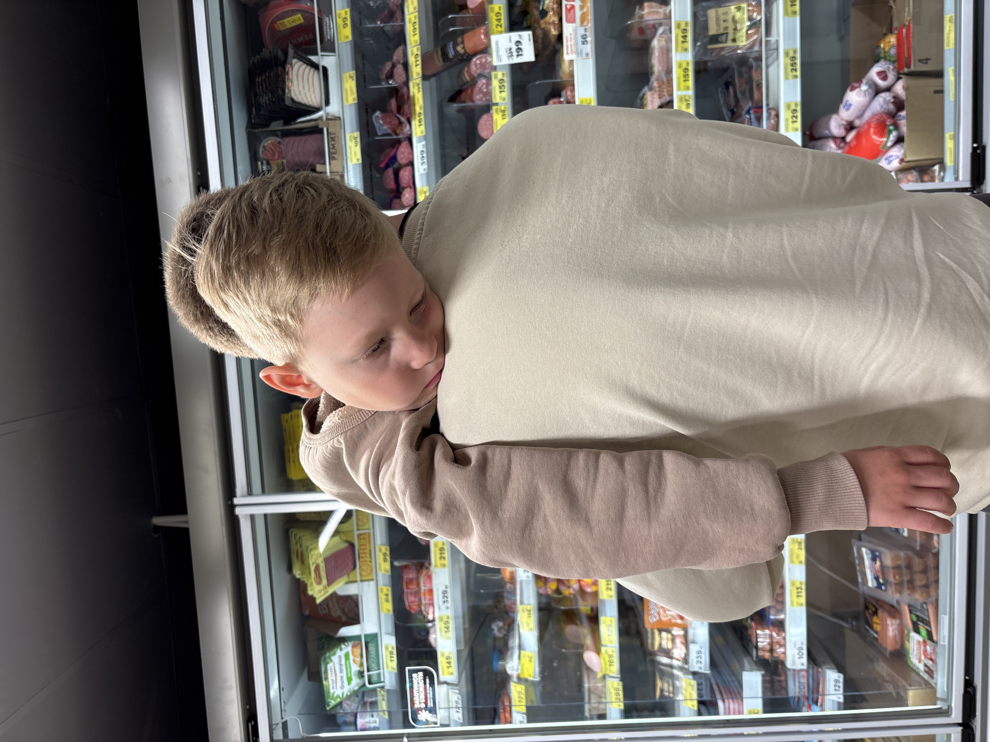 A young boy, appearing to be asleep or resting, is held over the shoulder of an adult. The boy has light brown hair and is wearing a light brown long-sleeved shirt. The adult is wearing a cream-colored sweatshirt. They are positioned in front of a refrigerated display case in what appears to be a grocery store. The display case is filled with various food products, predominantly meats and sausages, with price tags visible. The environment suggests a commercial setting within the city of Chaikovsky, Russia, likely a supermarket or a food market.