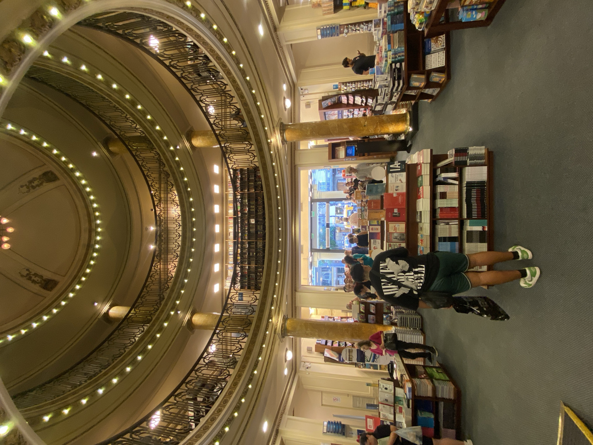 The image depicts the interior of El Ateneo Grand Splendid, a multi-level bookstore located in the Recoleta neighborhood of Buenos Aires, Argentina.
The scene captures the ground floor of what was formerly an opulent theater, characterized by a grand, curved ceiling adorned with multiple rows of small, warm lights and decorative molding. Several upper levels feature curved balconies with intricate black metal railings and gold embellishments. Large, fluted, marbelized columns with gold accents support the structure. The flooring is a dark grey/blue carpet.
Approximately 15-20 individuals are present on the ground floor, engaged in browsing books or walking through aisles. In the immediate foreground, with his back to the camera, is a male wearing a black t-shirt with a white graphic, dark green shorts, and white-and-green sneakers, holding a black shopping bag. Other individuals are seen interacting with book displays; for example, a woman in a pink top stands near a central column, and a man in a black t-shirt is visible to the right, looking at a mobile device.
Numerous books are arranged on dark wooden display tables and floor-standing shelves throughout the ground level. Some displays also include boxed retail items. In the background, a large doorway opens to reveal a street view, indicating the building's exterior and urban environment. Security cameras are visible mounted on columns.