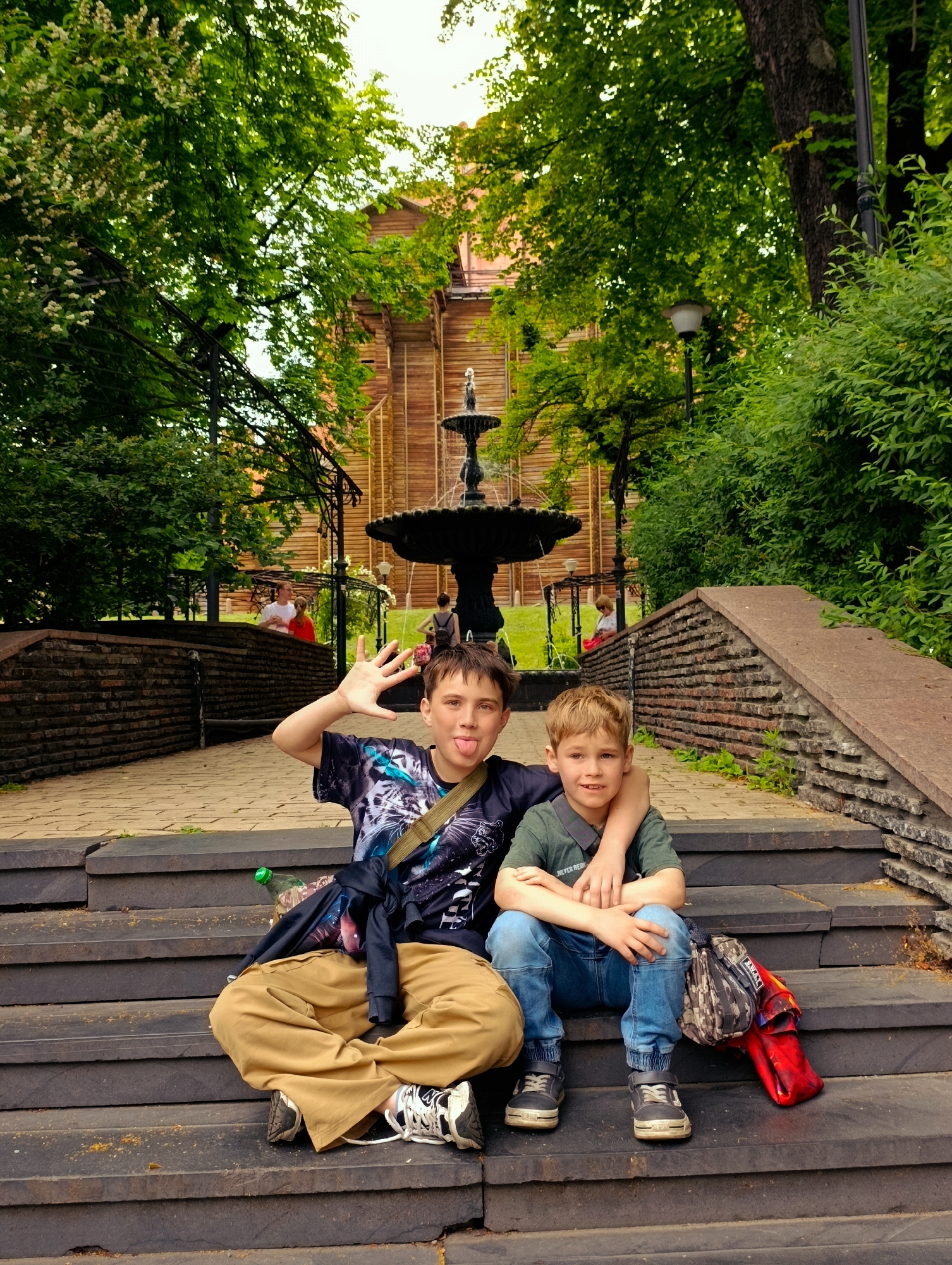 Two young boys are sitting on dark stone steps in an outdoor park. The boy on the left, who appears older, is wearing a black t-shirt with a graphic print, tan pants, and black and white sneakers. He is sticking his tongue out and holding up his left hand with fingers spread. The younger boy on the right is wearing a green polo shirt and blue jeans. He has his arm around the older boy. Both boys are looking towards the camera. A dark bag with a camouflage pattern and a red item are placed on the steps to the right of the younger boy. In the background, there is a multi-tiered fountain, a wooden building with a sloped roof, lush green trees, and other people walking. The scene appears to be in the Pechersk District of Kyiv, Ukraine, likely near the Kyiv Pechersk Lavra monastery complex.