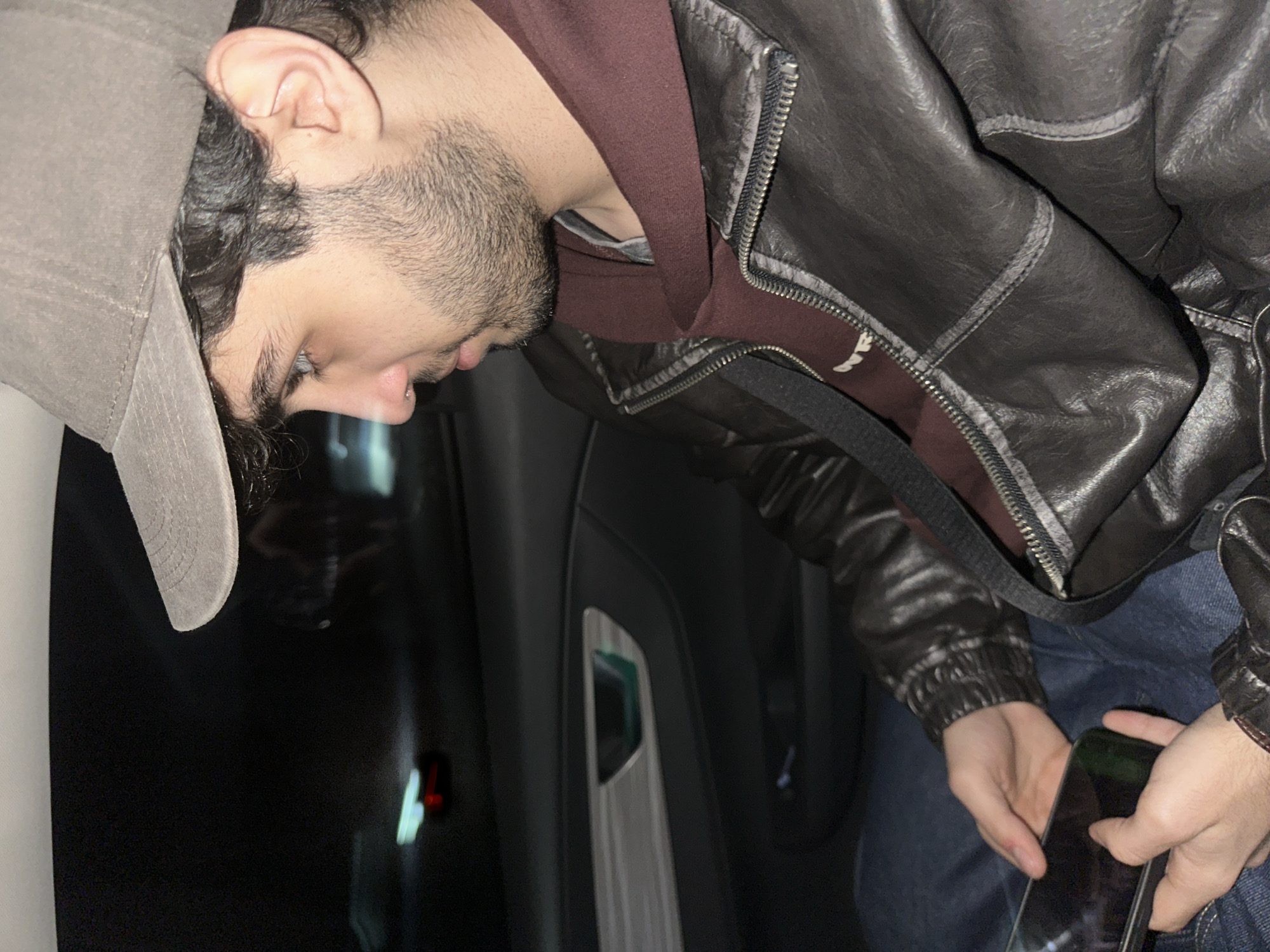 A man wearing a gray cap, a burgundy hoodie, and a dark leather jacket is seated inside what appears to be a car. He is holding a mobile phone in his hands. The interior of the car is dark, with a silver accent on the door panel visible. There is a red light on the door panel as well. The background is out of focus. There is no information available in the image that can confirm the location is Yerevan, Armenia.