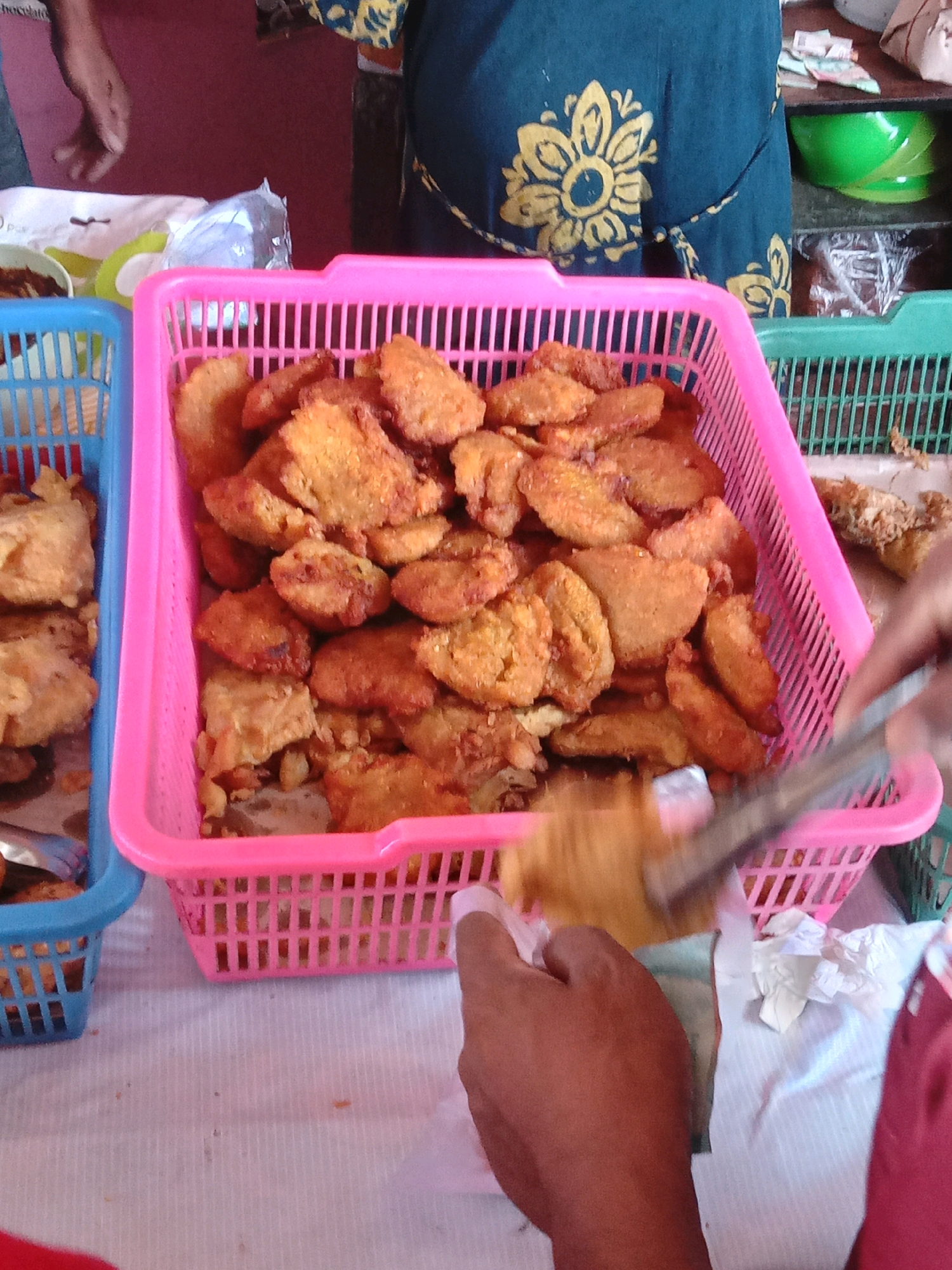 The image shows a street food stall in Surabaya, Indonesia. The stall is filled with baskets of fried bananas. A person's hand is reaching into a pink basket to pick up a piece of fried banana with tongs. There are other baskets of food and a green bowl visible in the background. The stall appears to be open-air.