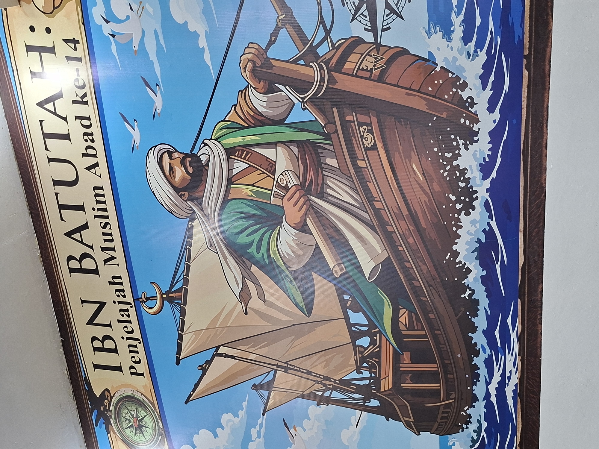 The image shows a framed illustration of Ibn Battuta, the 14th-century Muslim explorer, on a ship in the sea. He is depicted holding scrolls and wearing a turban and green robe. The ship is sailing through blue water with white waves and seagulls flying in the blue sky. The image is likely found in Lhokseumawe, Indonesia, as the title "IBN BATUTA: Penjelajah Muslim Abad ke-14" is in Indonesian, and the context suggests this is the case. The illustration is in a cartoon style, portraying Ibn Battuta as a heroic figure. A compass illustration is on the bottom-left of the image.