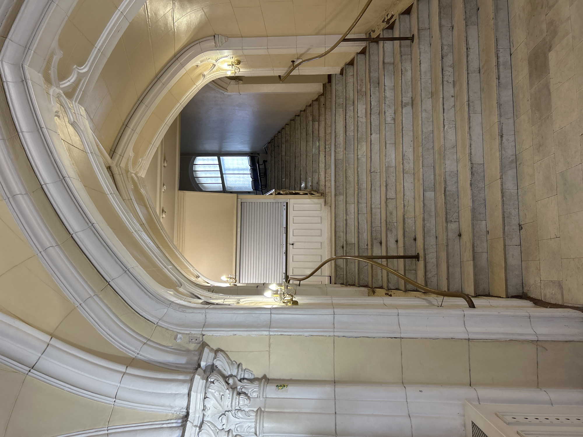 An indoor, ground-level, wide-angle shot captures a grand, curved staircase with marble steps and ornate, white-trimmed arches overhead. The staircase ascends towards a landing where a large, arched window with segmented panes illuminates the space. A metal security gate or shutter is partially visible on the left side of the landing, above a decorative white panel. Two ornate brass sconces with multiple bulbs are mounted on the wall near the staircase. A thin, brass handrail runs along the right side of the stairs. The walls are a pale cream color, and the architectural details suggest a historical or gothic style. The scene is well-lit, primarily by the light from the window and the sconces. There are no people or animals visible in the image.