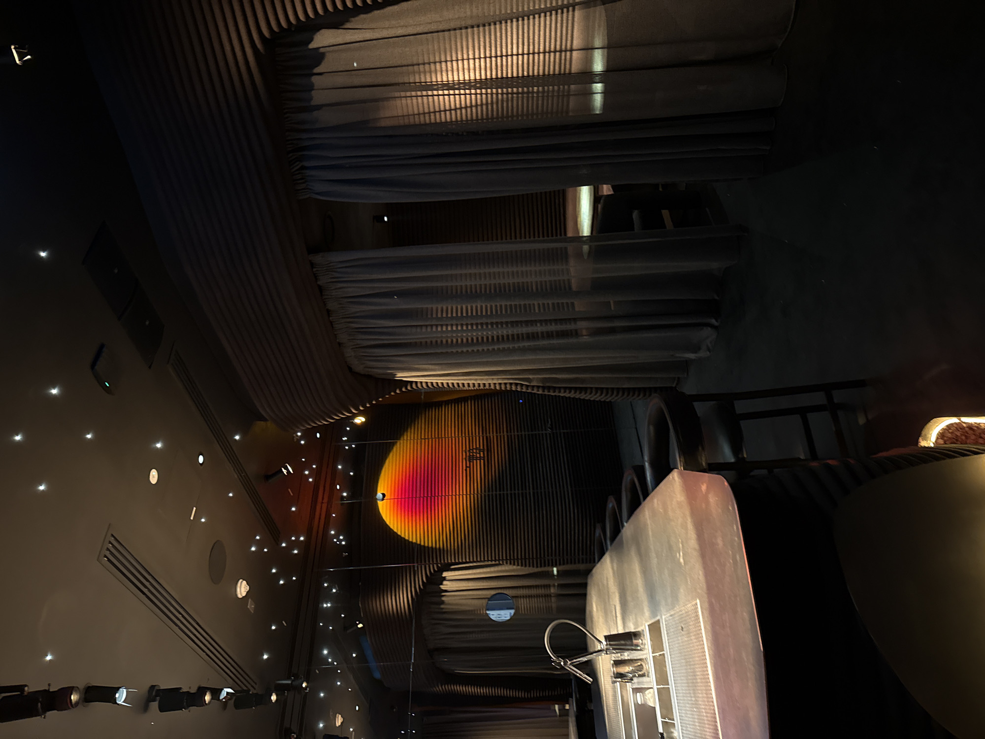 The image shows the interior of a restaurant or bar in Dubai, United Arab Emirates, characterized by its modern design and dim lighting. The ceiling features corrugated panels interspersed with rows of recessed lighting. Sheer curtains drape along sections of the ceiling. A wall is adorned with small, star-like lights. There is a bar counter, complete with barstools and equipment. A circular lighting installation with red, orange, and yellow hues is visible. The image is taken from an unusual angle, tilted upwards, emphasizing the ceiling and lighting design.