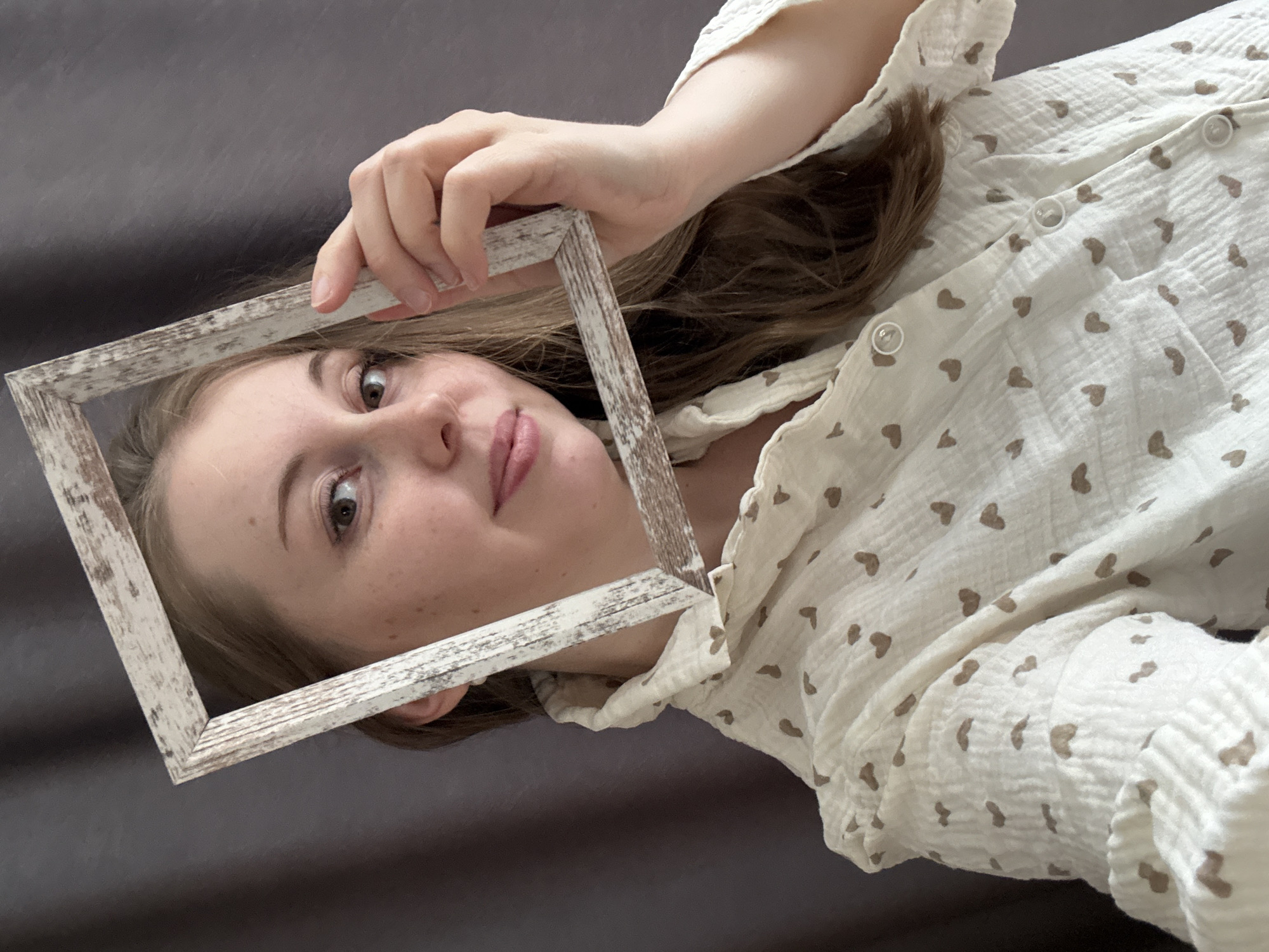 A young woman is captured in a close-up, angled shot, holding a distressed white wooden frame that partially encircles her face. Her head is tilted, and she looks directly at the viewer with wide, curious brown eyes. Her lips are slightly parted, revealing a subtle expression. Long, brown hair frames her face and shoulders.
She is wearing a light-colored (likely off-white or cream) shirt adorned with numerous small, brown heart-shaped patterns. The fabric appears soft and textured, possibly gauze or muslin, with visible buttons down the front. Her fair skin shows a scattering of freckles on her cheek. The background is a plain, dark gray or charcoal fabric, suggesting an indoor, casual setting, possibly lying on a bed or sofa. No text is visible in the image.