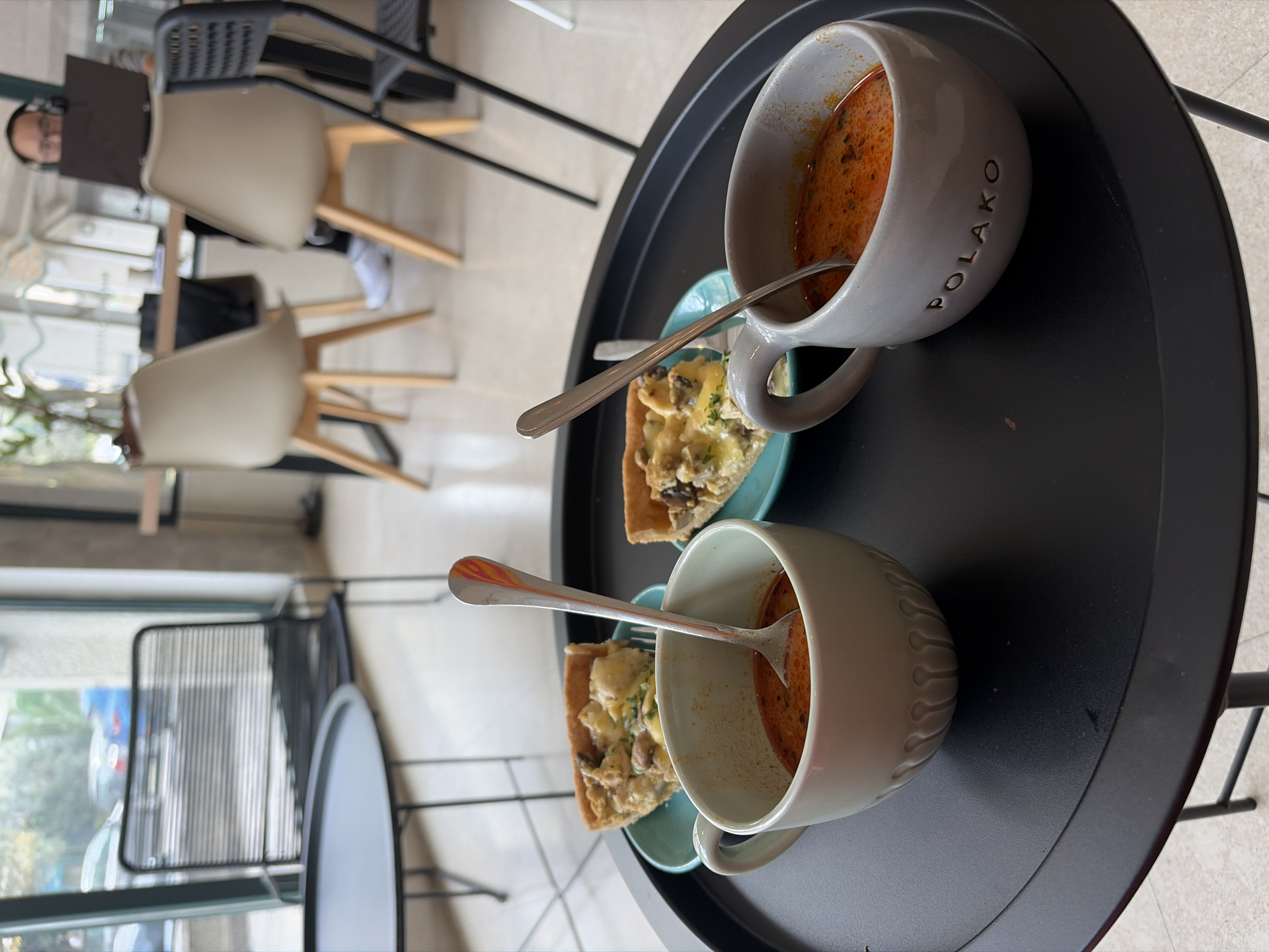The image shows a black round table in an indoor setting, likely a cafe or restaurant in Budva, Montenegro. On the table, there are two mugs filled with a reddish-orange soup. One mug has the word "POLAKO" inscribed on it. Beside each mug, there's a slice of quiche or a similar savory pie on a light blue plate, and a spoon is placed next to the quiche slice, partially extending into the soup mug.
In the background, there are chairs and tables, a person using a laptop, and a window with a partial view of the outside, potentially showing elements of the Budva cityscape. The scene suggests a meal or snack being enjoyed.