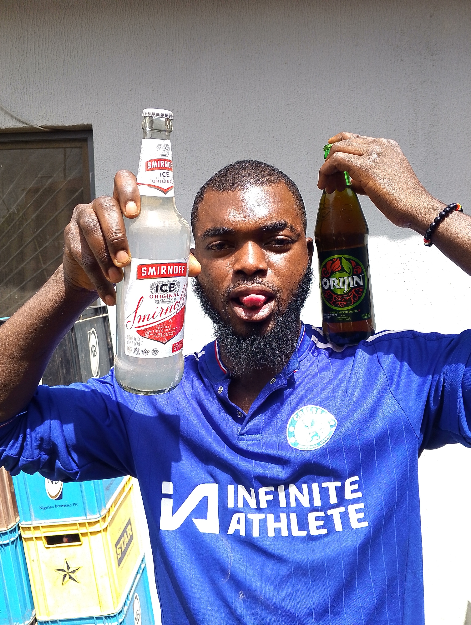 This is a medium shot, outdoor photograph of a young Black man in Jos, Nigeria. He is wearing a blue collared shirt with white pinstripes and has a logo on the left chest that appears to be a sports team crest. The text "INFINITE ATHLETE" is visible in large white letters across the front of his shirt.
The man is holding two bottles of alcoholic beverages. In his left hand, he holds a clear bottle of "SMIRNOFF ICE ORIGINAL". The bottle's label is predominantly white with red accents and features the Smirnoff logo. In his right hand, he holds a green glass bottle of "ORIJIN", a spirit mixed drink with a label featuring green and orange tones and images of fruit. He is looking towards the camera with his tongue sticking out playfully. He has a beard and is wearing a dark beaded bracelet on his right wrist.
Behind the man, there is a white textured wall. To his left, a stack of blue and yellow plastic crates is visible. Some of the crates have text printed on them, including "Nigerian Breweries Pic", "NB", and "STAR". The lighting suggests it is daytime, likely sunny given the bright illumination and shadows. The overall scene conveys a casual and fun atmosphere, possibly a moment of enjoyment or celebration.