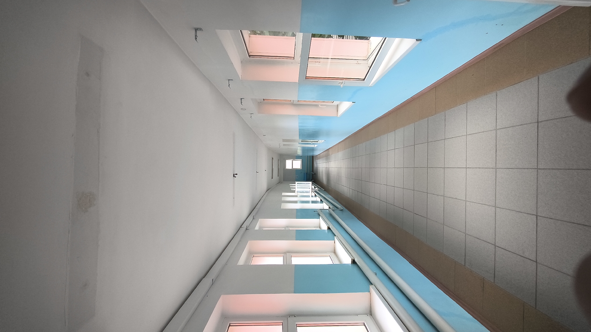 A long, well-lit hallway with blue and white walls and a tiled floor. There are windows on both sides of the hallway, letting in natural light. The hallway appears to be empty, with no people or objects present. The ceiling has a patch that looks like a repair.