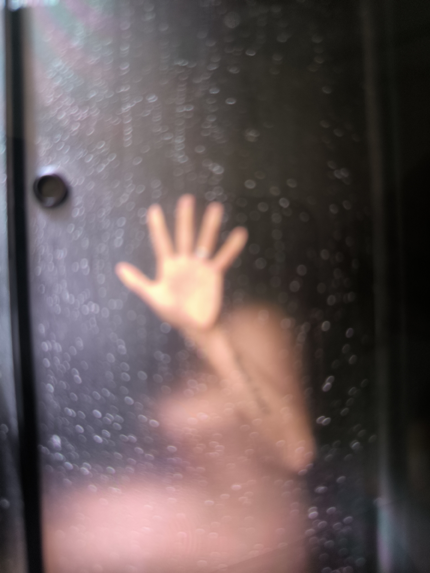 A blurry image shows a hand pressed against a steamy glass surface, obscuring a partial view of a person's silhouette. The environment appears to be inside a shower stall with a dark interior. The location of Tbilisi, Georgia, is not discernible from the image.