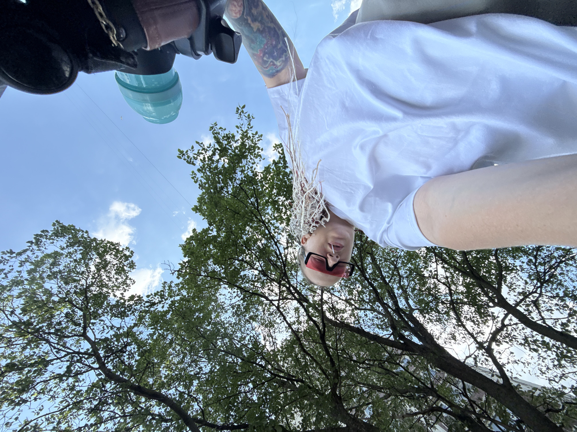 A close-up, low-angle, upside-down selfie shows a person wearing a white t-shirt and red-tinted sunglasses. The person has a shaved head with thin, blonde braids. A tattoo is visible on their left arm. The background is a bright blue sky with scattered clouds and the leafy branches of trees. In the foreground, parts of what appear to be a bicycle or scooter are visible, including a black handlebar, a chain, and a turquoise-colored accessory. The scene is outdoors, likely in a park or street in Moscow, Russia, given the foliage and architecture suggested by the building in the upper right corner.