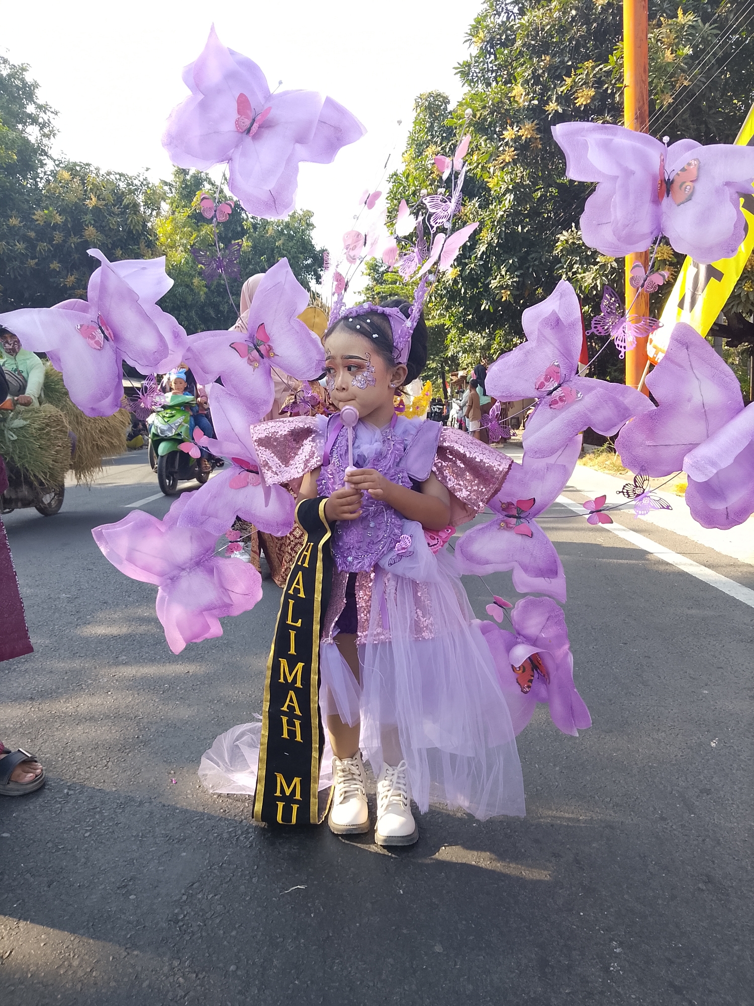The image depicts a young girl participating in a parade, likely in Buduan, Indonesia, given the visual context. She is adorned in an elaborate purple costume designed to resemble a butterfly, complete with large, translucent purple wings that extend from her back. The costume is decorated with smaller pink butterflies and sequins. She has face paint, including glitter on her cheeks and eyelids, and wears a purple headband with butterfly accents. In her right hand, she holds a pink lollipop. A black sash with gold lettering, reading "HALIMAH MU", is draped across her body. She is wearing white lace-up boots.
The parade is taking place on an asphalt road, with trees and vegetation visible in the background. Other participants and spectators are present, including a person on a green motorcycle and a cart laden with hay. The lighting suggests it is daytime, likely with bright sunlight creating shadows on the road. The overall scene captures a festive and celebratory atmosphere typical of a cultural event or parade.