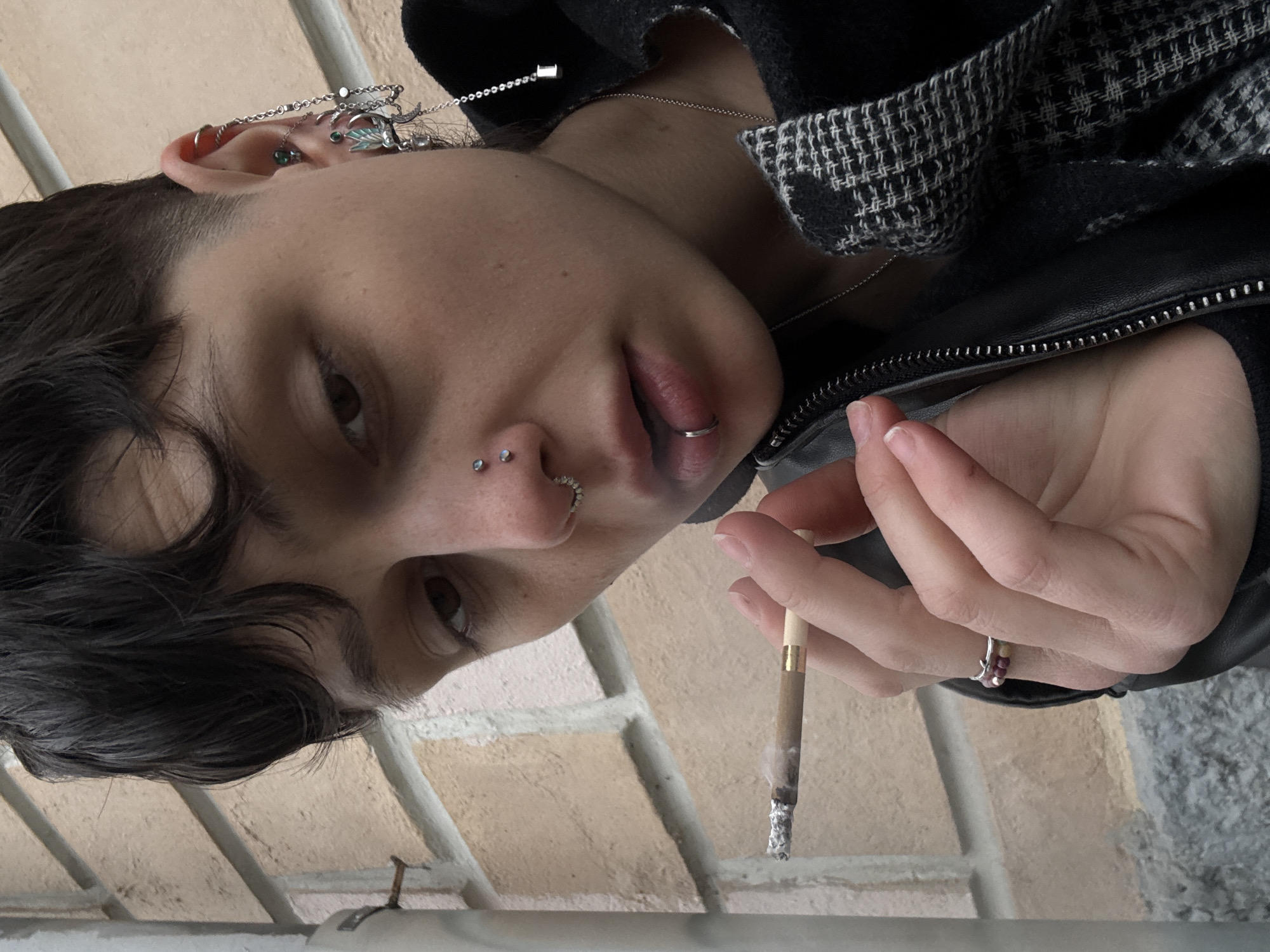 A close-up, slightly angled shot shows a person with short, dark hair holding a lit cigarette. The person has several piercings: two in their left nostril, one on their lower lip, and multiple piercings in their left ear adorned with chain-like jewelry. They are wearing a black leather jacket over a black and white patterned garment. A ring is visible on one of their fingers. The background features a beige brick wall and a gray surface, suggesting an outdoor setting. The image does not reveal any information to specifically link the image to the city of Moscow, Russia.
