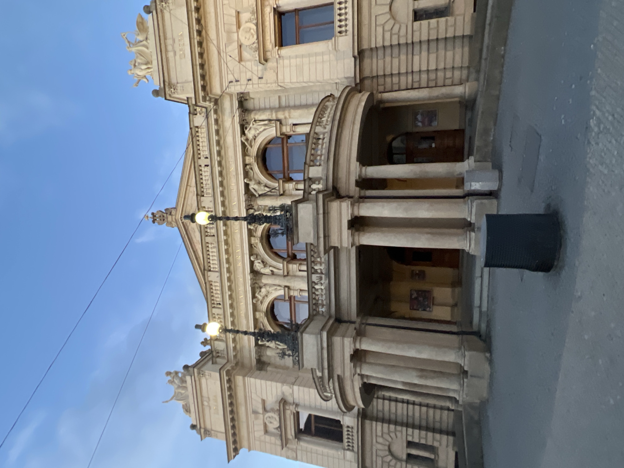 The image shows the facade of the New Theatre in Pilsen, Czechia. The theatre is a large, ornate building with classical architectural elements. It features a prominent portico with tall columns supporting a balcony. Above the portico, there are arched windows and decorative sculptures. The building is made of light-colored stone and appears to be illuminated by streetlights, suggesting it is either dusk or dawn. The sky above is blue with some wispy clouds. In the foreground, there is a paved area with a dark cylindrical trash receptacle and a lighter, rectangular trash receptacle. There are also power lines running across the sky. The overall scene depicts a public building in an urban setting.