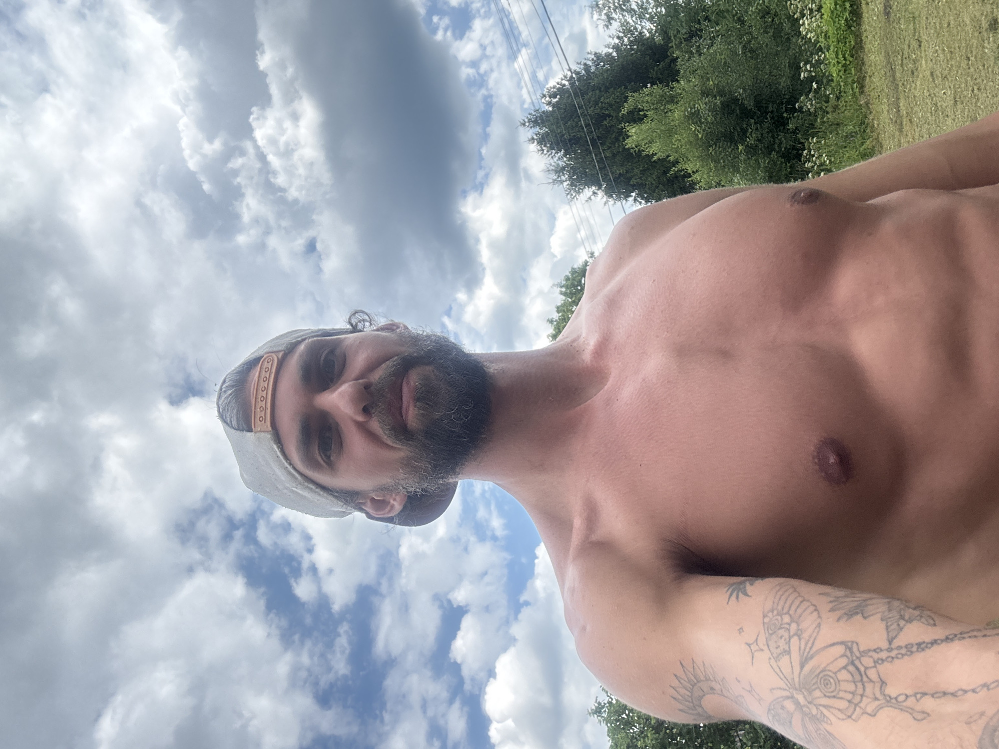 This media file is a selfie taken outdoors under a cloudy sky. The primary subject is a shirtless man with a beard, wearing a baseball cap. He has tattoos on his arm, including a butterfly design. The background shows greenery, suggesting a rural or natural setting, possibly in Kuti, Russia. The lighting and clouds indicate it is daytime, likely overcast or with intermittent sun. The man's facial expression is neutral, and the overall mood is relaxed and casual. There is no visible text in the image.