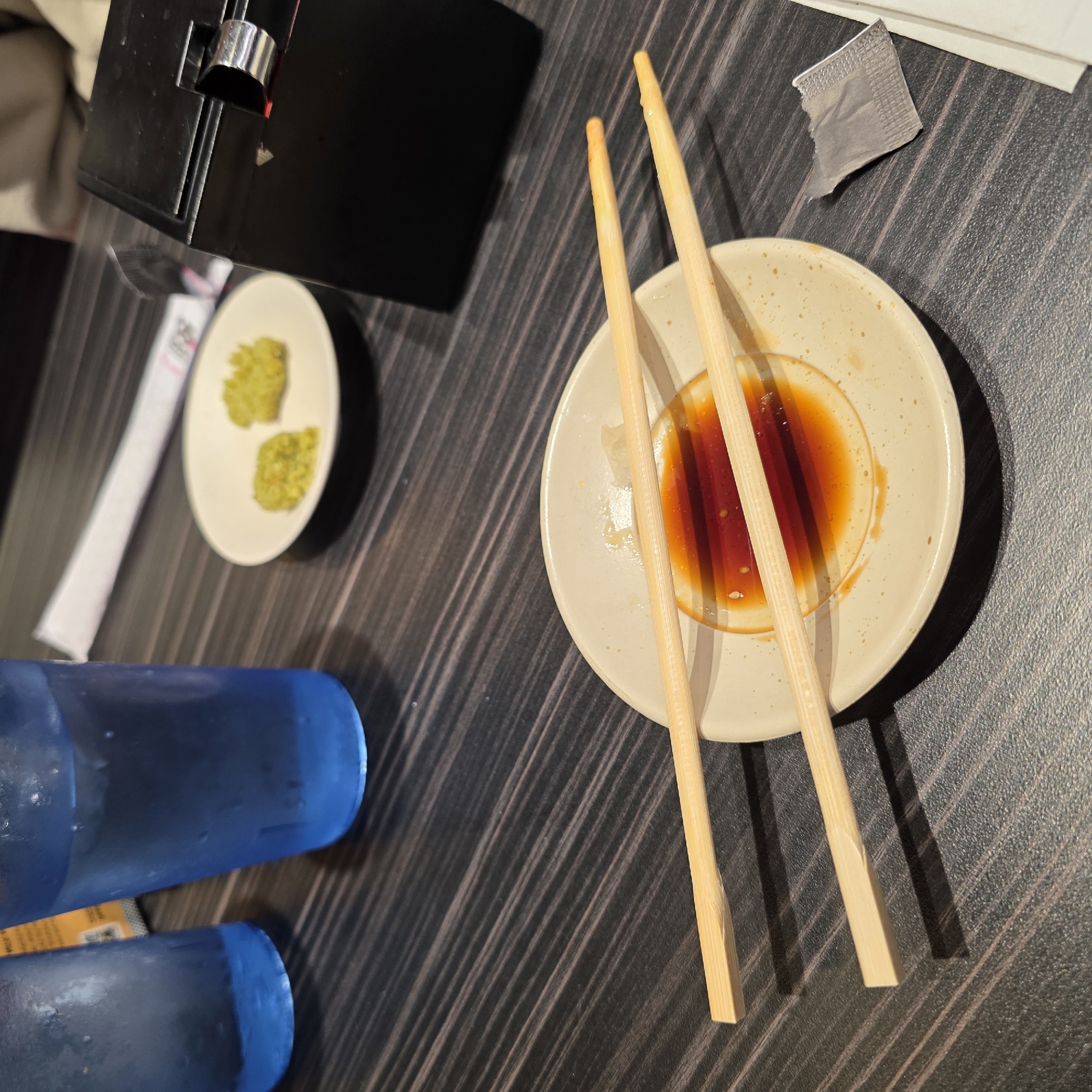 A close-up, eye-level shot shows a dark wood grain table with two blue glasses of water and a black napkin holder on the upper right. In the center of the frame, a light-colored plate holds a small amount of soy sauce and two wooden chopsticks resting on top, partially submerged in the sauce. To the left of the plate, two small mounds of green paste are on a smaller white dish. A white paper wrapper lies near the bottom right of the frame. The scene appears to be in a restaurant in Beaverton, Oregon.