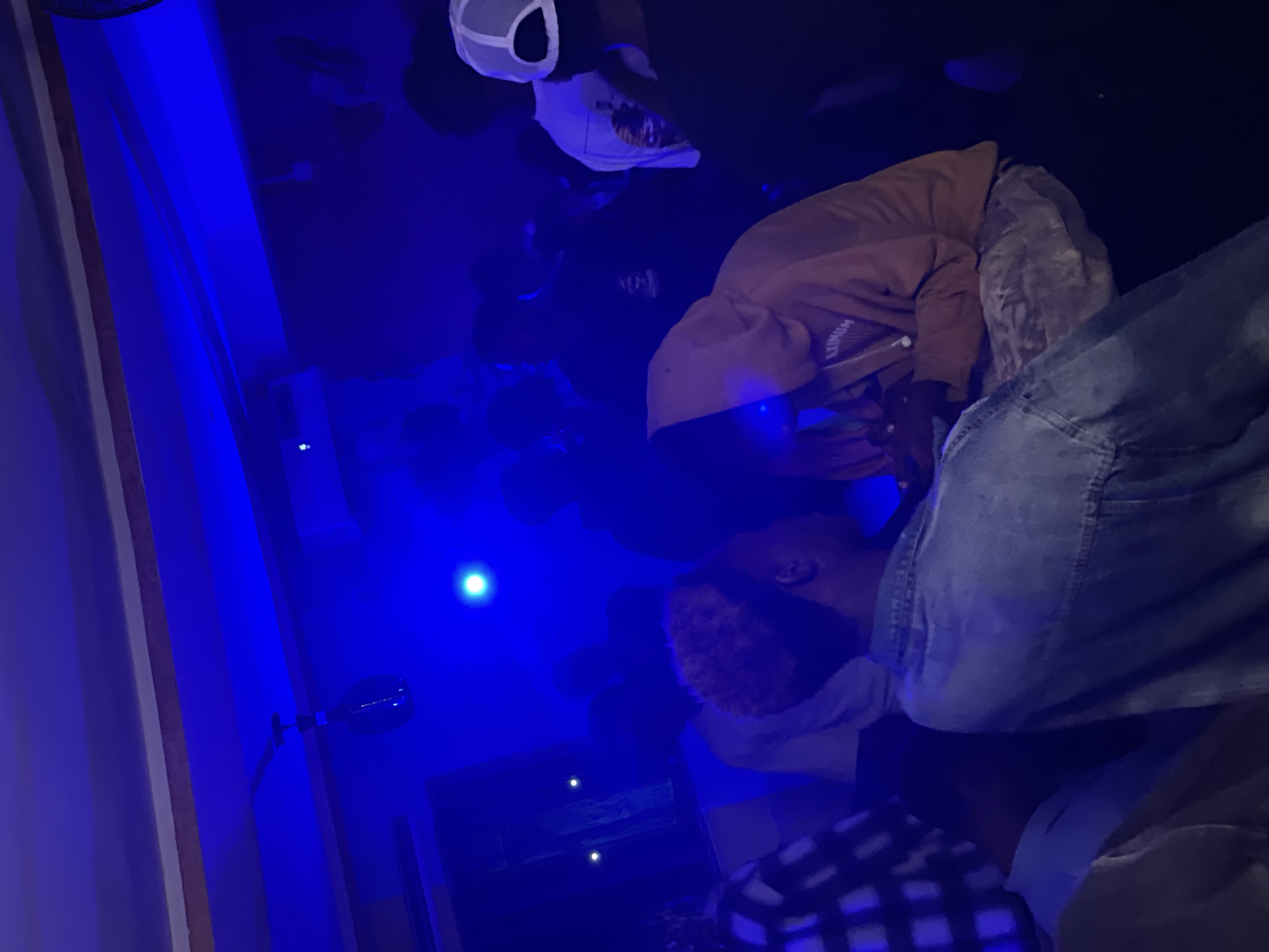 The image depicts an indoor gathering, likely a party or social event, illuminated by predominantly blue lighting. Several individuals are present, with varying degrees of visibility due to the low light conditions and their positions within the frame. In the foreground, a person in a denim jacket is seen from the back, facing away from the camera, with their head turned to their left. To their right, another individual wearing a tan hoodie is partially visible, with their head lowered, appearing to be engaged with something in their hands. Behind these individuals, a crowd of people are gathered, their features mostly obscured by shadow and the blue light. Some individuals are wearing hats, including a white baseball cap. The environment suggests a dimly lit indoor space, possibly a club or a private residence, with what appears to be a ceiling fan and a light fixture visible in the background. The overall atmosphere is that of a social gathering in the city of Durumi, Nigeria.