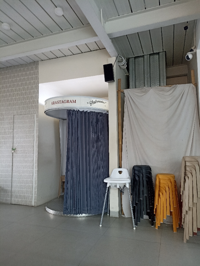 The image shows an indoor photo studio with a sign that reads "ARASTAGRAM - Photomatics." The studio is located in Purwokerto, Indonesia (latitude: -7.42, longitude: 109.245). The studio has gray tiled floors, white walls with a textured pattern, and a white ceiling with wooden beams. There is a black curtain in a photo booth behind a circular white platform. To the right of the photo booth is a white sheet hanging from the ceiling. There are three chairs in front of the sheet. One chair is white, one is black, and one is yellow. The chairs are in front of a white wall with a textured pattern. There is a speaker on the wall to the left of the sheet. There is a white door to the left of the photo booth. The studio appears to be empty and unused.