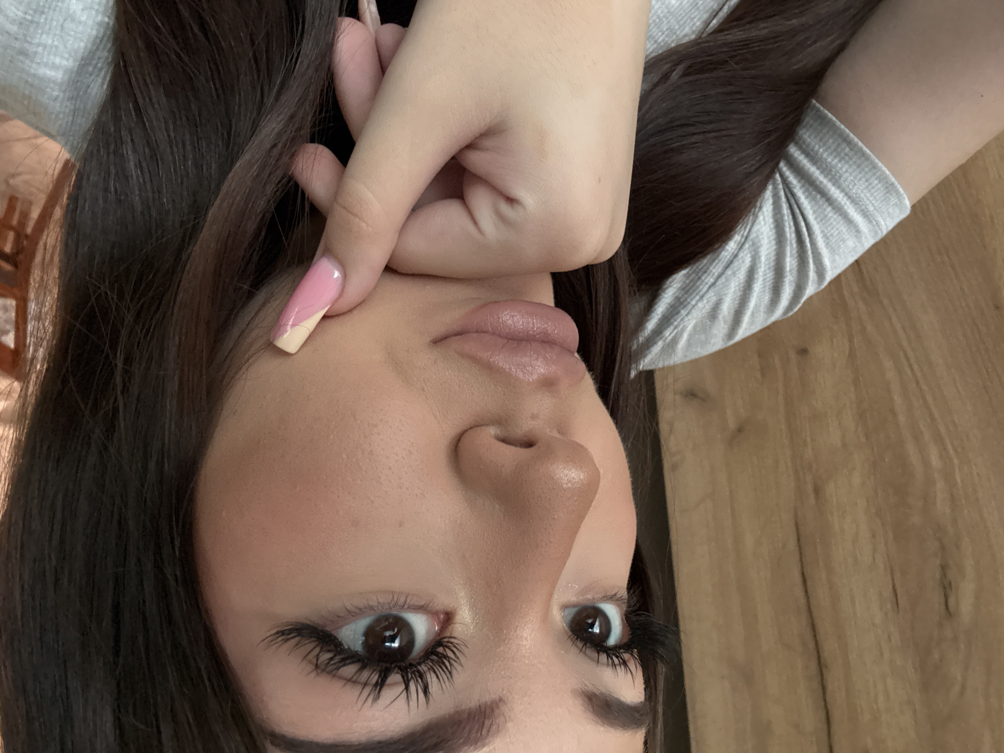 A selfie taken from a low angle of a woman with long brown hair, wearing a light gray long-sleeved shirt. Her face is the primary focus, with her brown eyes looking directly at the camera. She has prominent false eyelashes and defined eyebrows. Her skin appears smooth and made up, with light pink lips. Her left hand is near her face, with a long nail painted pink and beige. The background is a wooden surface, possibly a table or floor. The location is not discernible from the image.