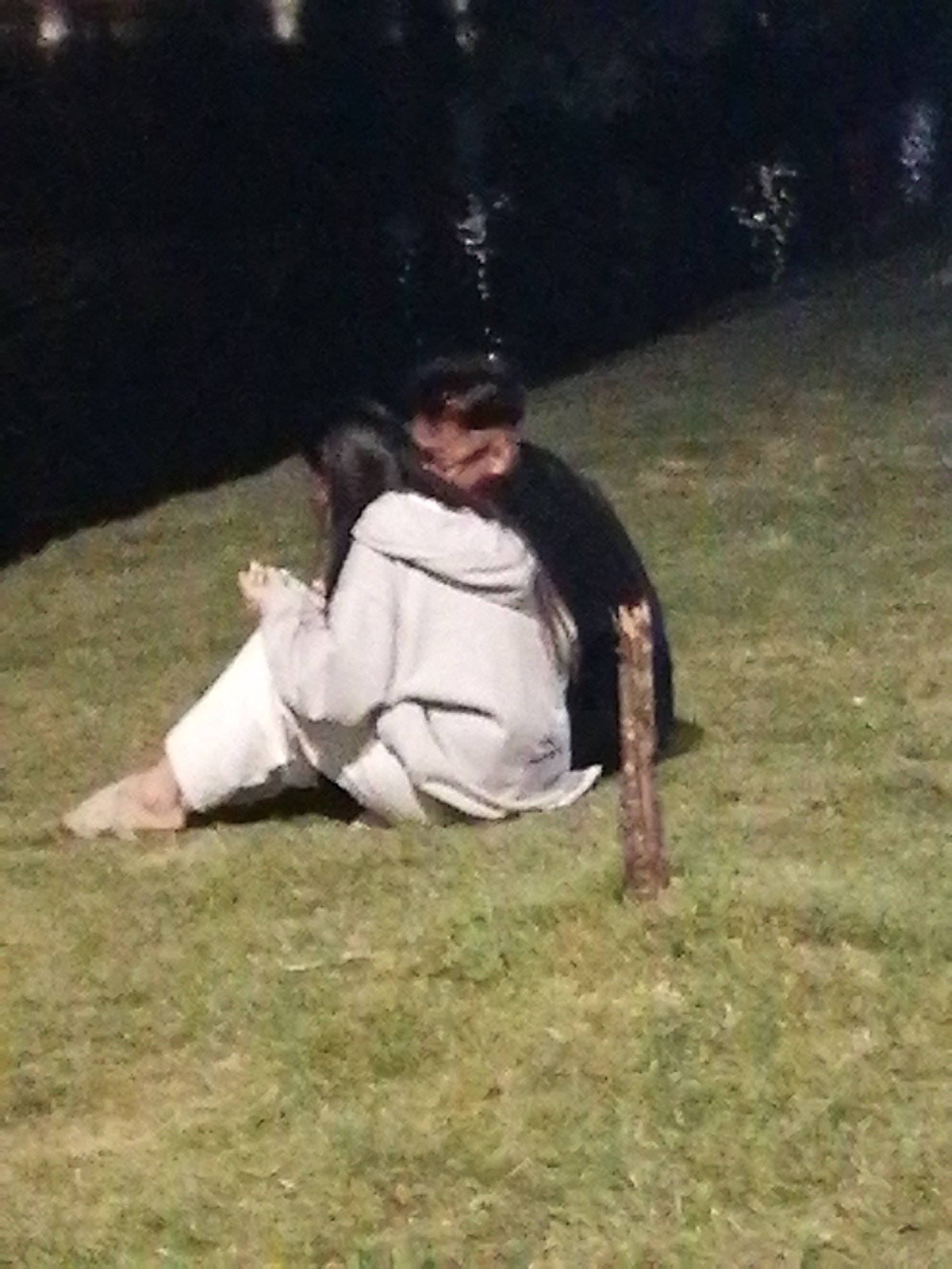 A dark, blurry image shows two people sitting on grass at night. The person on the left is wearing white pants and a light-colored hooded sweatshirt. They are sitting with their legs tucked underneath them, facing to the left. The person on the right is wearing dark clothing and has their arm around the person on the left. They appear to be leaning in. A wooden object, possibly a piece of driftwood, stands vertically in the grass to the right of the couple. The background is dark, with some out-of-focus lights visible, suggesting a dimly lit outdoor environment. The image is grainy and lacks sharp detail, making it difficult to discern specific features of the individuals or their surroundings. It is not possible to determine the location within Da Lat, Vietnam, based on this image.