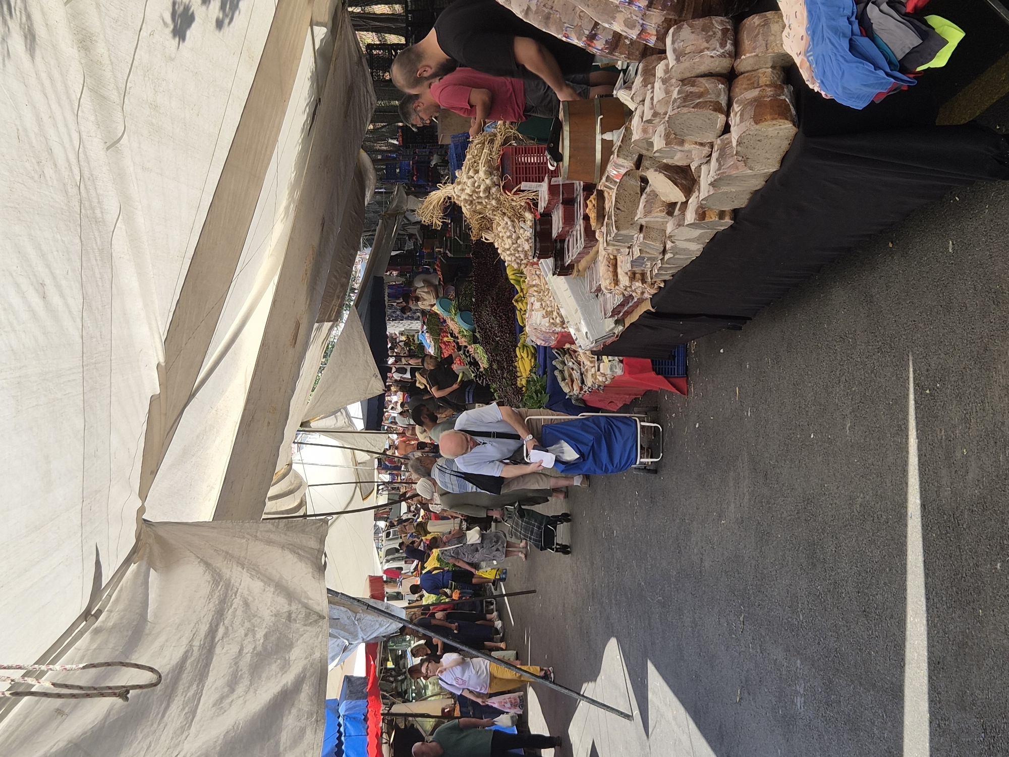 This is a vertical, eye-level, medium shot of a busy outdoor market in Maltepe, Turkey, on a sunny day. The market is covered by a large, off-white tarp, casting shadows on the asphalt ground. A variety of vendors are selling their goods, with people browsing and shopping.
In the foreground, a table is covered with a black cloth and laden with various food items, including loaves of bread, jars, and what appear to be small containers of preserves. To the right, a stack of folded clothing is visible.
Further into the market, a man in a light blue shirt is pushing a blue shopping cart. He is looking down at the cart as he walks. Behind him, a woman in a grey dress is pulling a checkered shopping cart. To the left, a woman in a white shirt and yellow pants is walking with a shopping bag in her hand. A man in a dark green shirt is walking on the far left.
The background reveals more market stalls with produce, including piles of dark berries, bananas, and onions. Several people are seen shopping and interacting with vendors. In the distance, vehicles and buildings are visible, suggesting an urban setting. The overall atmosphere is one of commerce and community activity.