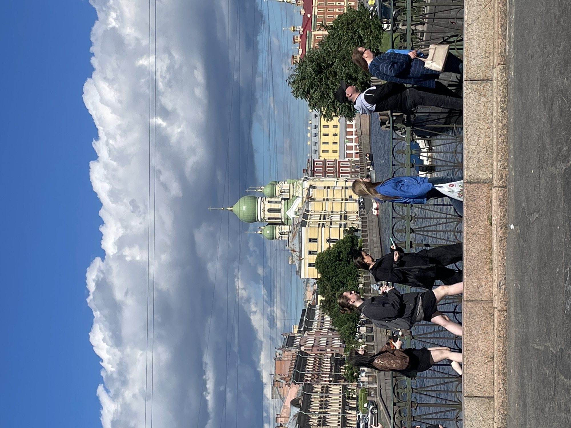 This image captures a group of people on a bridge overlooking a cityscape. The scene suggests a pleasant day with a mix of bright blue sky and dramatic, dark clouds, hinting at changing weather. The time of day appears to be daytime, likely late morning or afternoon, given the lighting.
The subjects are several individuals positioned along the railing of the bridge. Some are looking out at the view, while others are engaged in conversation or using their phones. The architecture of the city visible in the background features buildings with a distinct European style, including domes that suggest a church or cathedral. The presence of a body of water, likely a river or canal, beneath the bridge and the detailed urban landscape further place the scene in a city setting, specifically Saint Petersburg, Russia, as indicated.
Notable details include the contrast between the fluffy white clouds and the darker, more ominous ones, creating visual interest in the sky. The people are dressed in casual attire, suggesting a relaxed atmosphere. One person is carrying a light-colored tote bag, and another has a dark backpack.
The overall impression is one of a dynamic urban environment on a day with varied weather conditions, with people enjoying the scenery and each other's company. There is no visible text in the image.