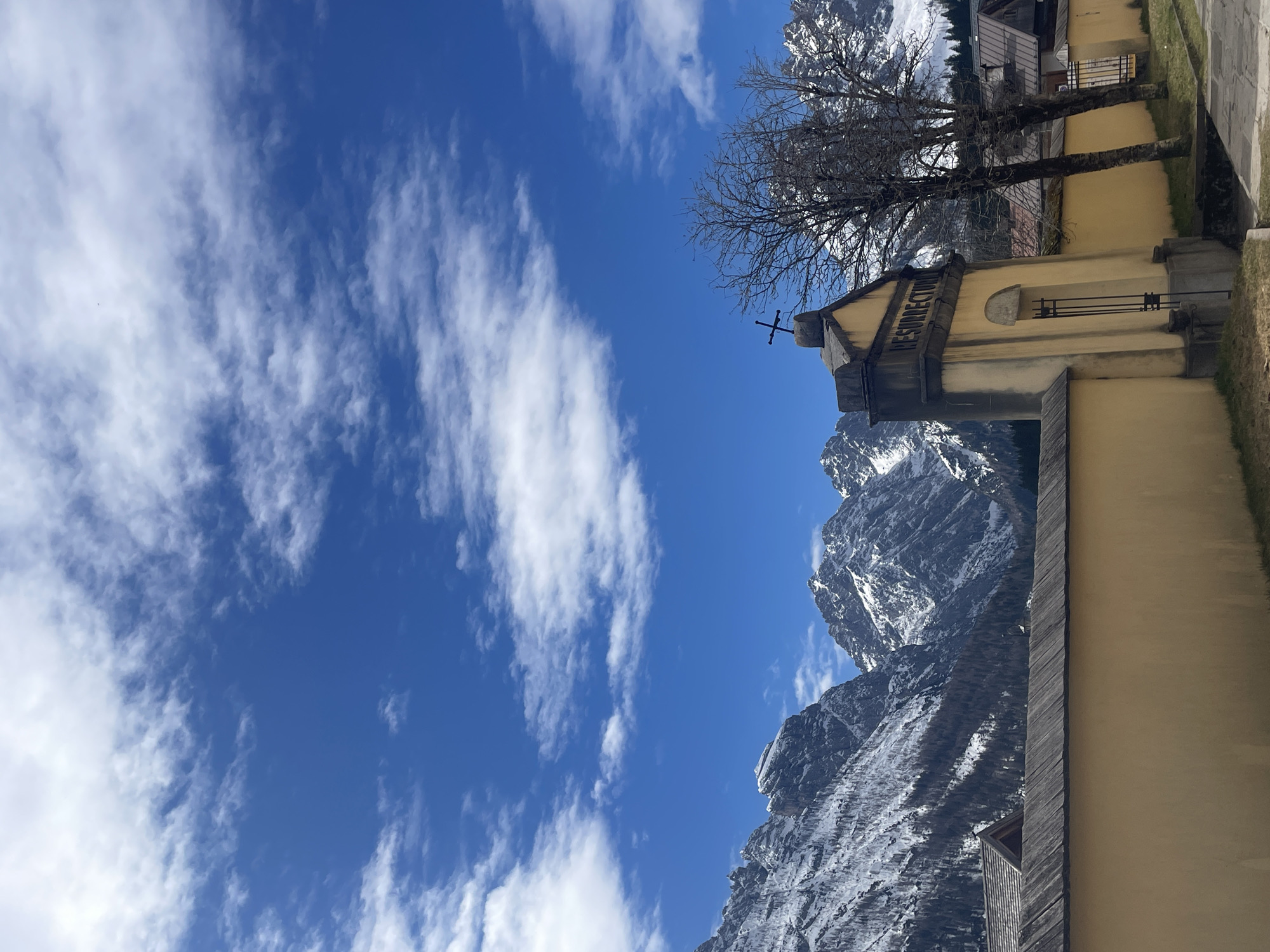 The image shows a view of a church bell tower in Sappada/Plodn/Sapade, Italy, set against a backdrop of snow-capped mountains and a blue sky with scattered clouds. The tower is a light yellow color, with a cross at its peak and the word "RESURRECTURUS" inscribed on its side. A bare tree stands nearby. Part of the adjacent building is visible, with some stone and wooden trim. The mountains rise in the background, partially covered with snow, and the sky is clear with wispy white clouds.