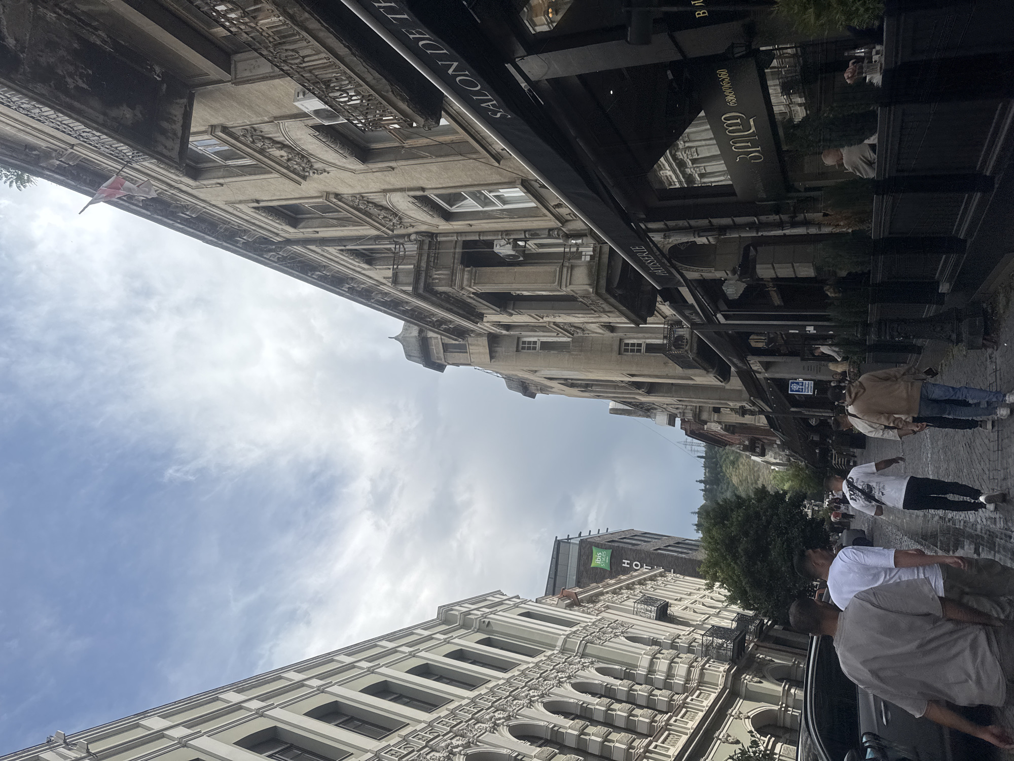 The image depicts a street scene in Tbilisi, Georgia. The architecture on both sides of the street is old and ornate, with balconies and detailed facades. On the right side of the street, there is a building with awnings that read "PATISSERIE" and "SALON DE THÉ". Further down the street on the right, another establishment has text in Georgian script visible. There is a flag with a red cross on a white background flying from the top of a building on the right. On the left side of the street, there is a building with arched windows and decorative elements. A sign for the "ibis STYLES HOTEL" is visible on a darker building behind the ornate facade.
Several people are walking down the street. In the foreground, four men are walking away from the camera, with one wearing a grey t-shirt, another in a white t-shirt, and the other two in casual clothing. Several other individuals are visible further down the street, some walking and others seated at outdoor tables. The street appears to be wet, suggesting it may have recently rained or is about to rain, as indicated by the overcast sky. Cars are parked along the left side of the street. The overall impression is of a historic urban environment.