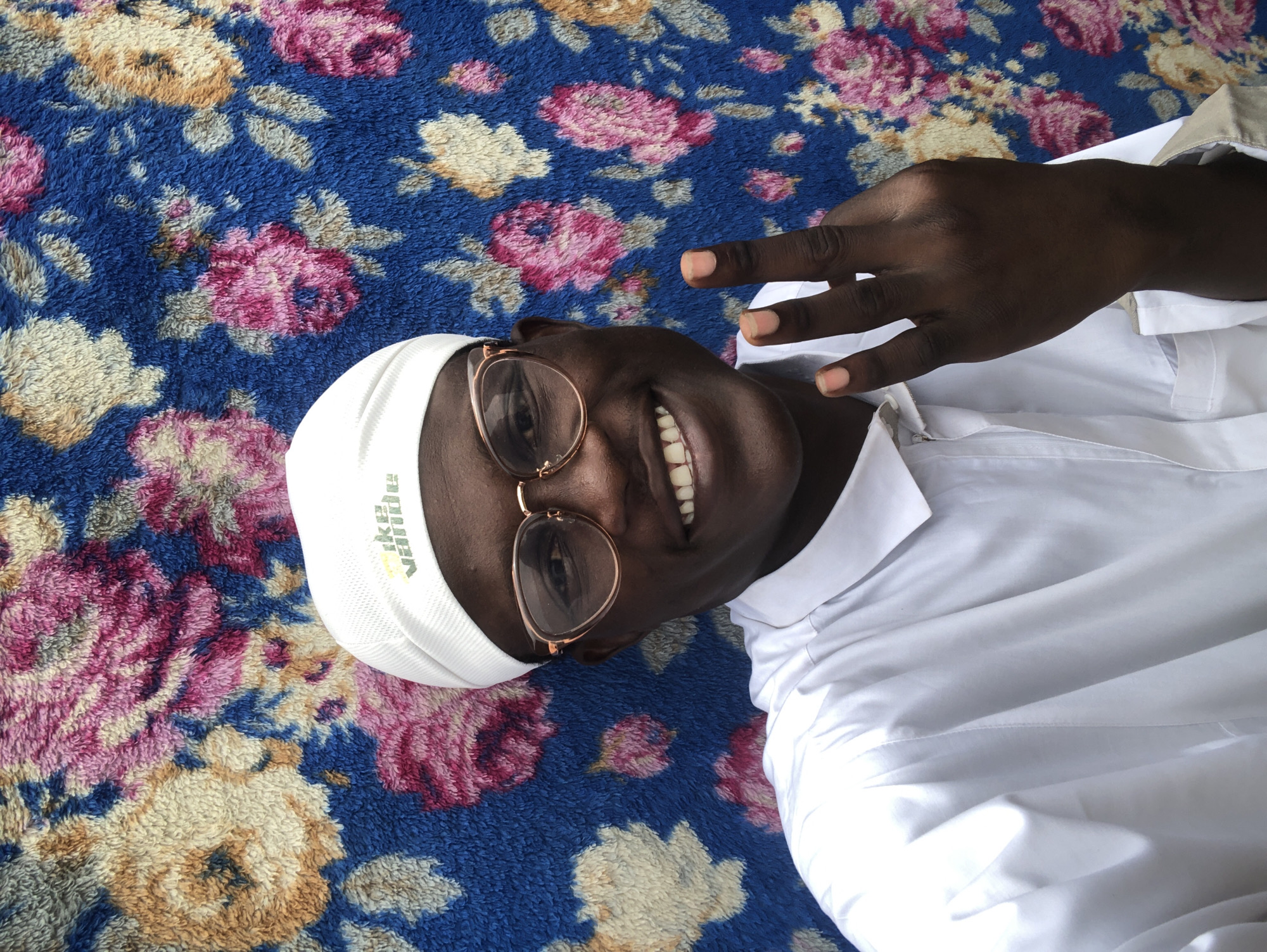 A dark-skinned young man wearing a white kufi and a white shirt, with round, light brown framed glasses, smiles at the camera and holds up two fingers in a peace sign. He stands in front of a bright blue, floral-patterned blanket or tapestry. The setting appears to be indoors, possibly a home, in Ado, Nigeria.