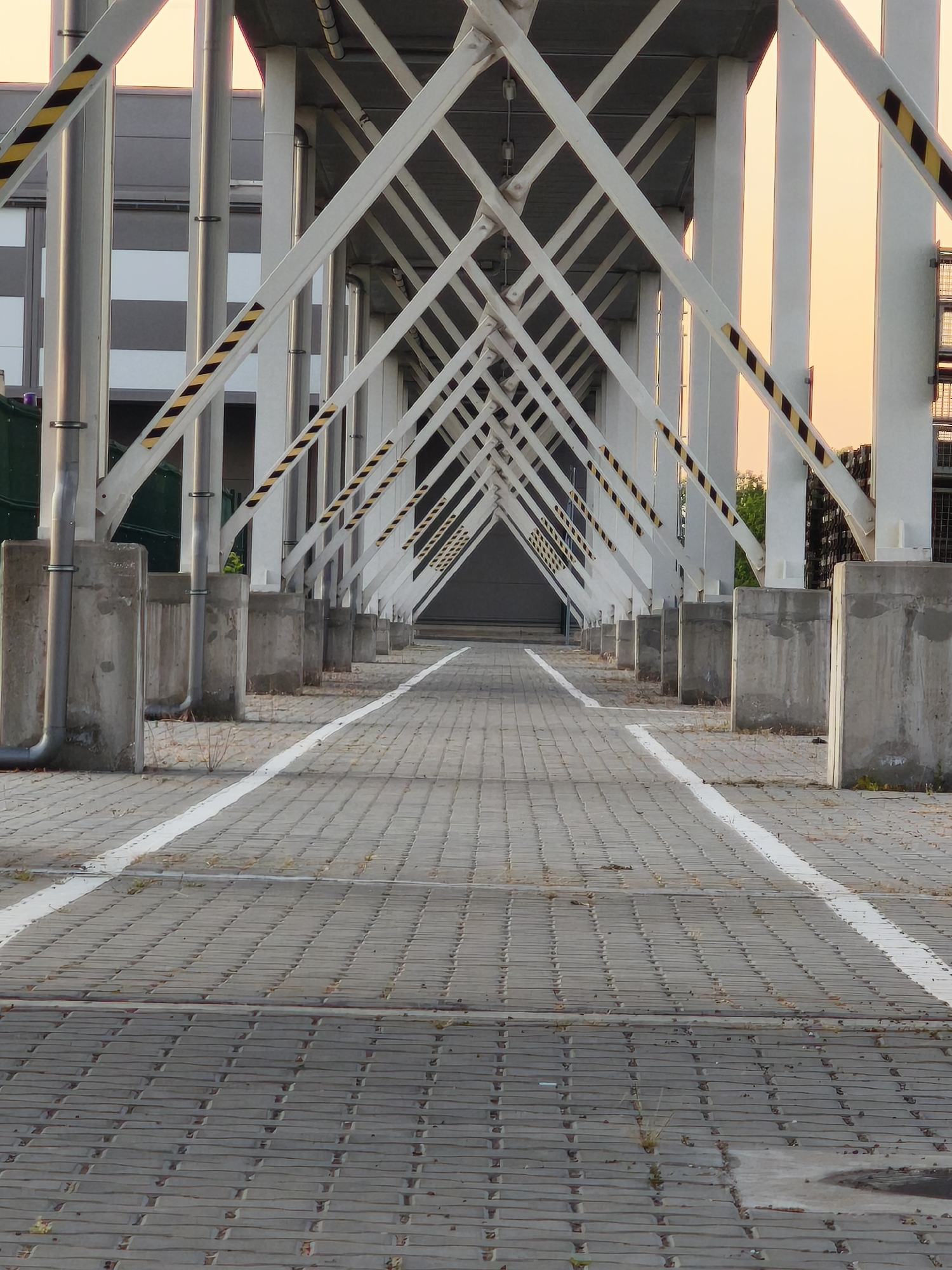 The photograph depicts a perspective view of an outdoor passageway in Benkovski, Bulgaria. The passageway is covered by a network of white steel beams arranged in a series of intersecting diagonal lines, supported by vertical steel columns. Each column is reinforced at its base by a concrete support block. Black and yellow hazard stripes are applied to the lower portions of the diagonal beams and some of the vertical columns. A paved walkway, delineated by parallel white lines, runs through the center of the passageway. The paving consists of interlocking gray concrete blocks. In the background, a building with gray and white striped paneling is visible on the left, while a structure with a grated metal staircase is on the right. The sky is visible at the far end of the passageway, exhibiting a pale orange hue. Small patches of vegetation are visible growing between the concrete paving blocks and around the base of the concrete supports. No people are present in the scene.