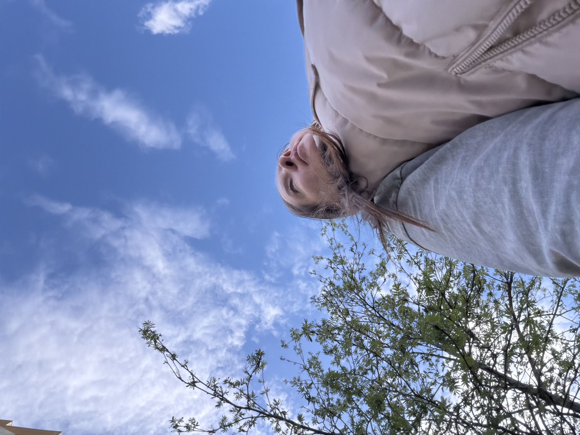 The photograph, taken from a low angle, features a woman looking upwards towards a blue sky with scattered white clouds. She is wearing a beige puffer jacket and a grey top. Part of a tree with green leaves and branches is visible in the lower right corner. A portion of a building can be seen in the bottom left corner. The location is presumed to be outdoors. The city of Staraya, Russia is specified as the location.