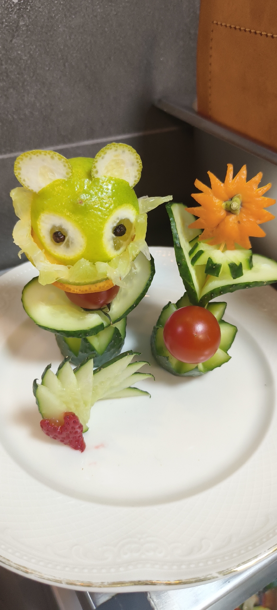 The image displays an artistic food presentation on a white plate with subtle embossed patterns and a gold rim. Two distinct, whimsical creations, primarily made from fruits and vegetables, are arranged on the plate.
On the left, a creature resembling a frog or bear forms the main focal point. Its head is crafted from a green citrus fruit, possibly a lime, with two small, dark seeds (likely peppercorns) set into carved-out sections to form eyes. Thin, semicircular slices of citrus peel act as ears, and a round slice of orange or mandarin forms its mouth. This head rests upon a stack of thick cucumber slices, which form the body. A single cherry tomato is positioned below the head, and further down, fanned-out cucumber slices suggest legs or a tail. A small, carved piece of strawberry, potentially shaped like a heart or foot, is at the very bottom of this structure.
To the right, a second, smaller decorative piece is present. It consists of geometrically cut and arranged cucumber slices that form a base. A bright orange, star-shaped or flower-shaped carving, possibly from a carrot or another citrus peel, is perched on top, held by a small toothpick. Another cherry tomato is nestled within the cucumber base of this arrangement.
The background features a dark grey, textured wall or surface on the left, and a warm brown object, possibly leather-bound furniture or a box, in the upper right corner. The lighting is bright and even, suggesting an indoor setting. There is no visible text. The overall presentation indicates a creative and playful approach to food preparation, possibly for a meal or as a decorative display.