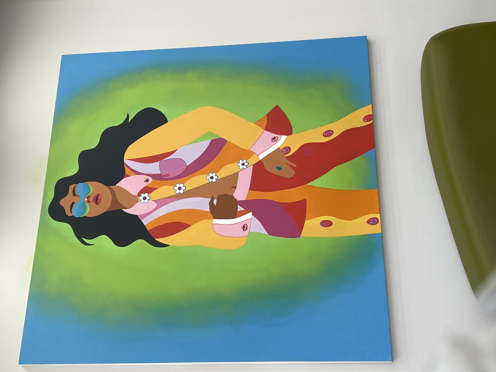 The image shows a colorful painting of a person wearing a vibrant suit. The person has dark hair and is wearing large blue sunglasses. The suit is a patchwork of yellow, orange, red, pink, and purple. There are small white flowers decorating the front of the suit and red circular details on the pants. The background of the painting is a gradient of bright green and blue. The painting is hung on a white wall, and a portion of a green chair is visible in the foreground. There is no indication of the location in the city of Yekaterinburg, Russia.