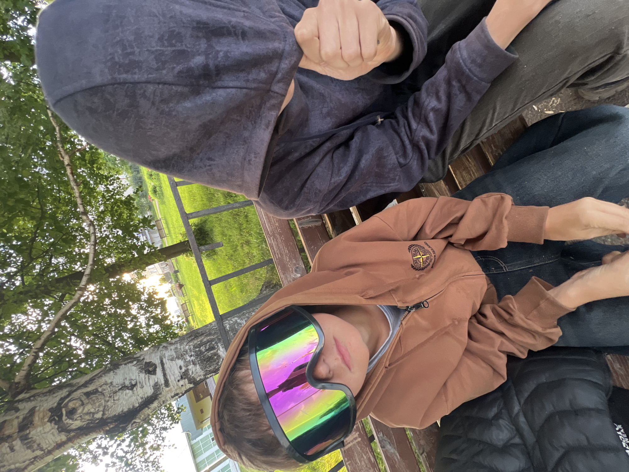 Two people are sitting on a wooden bench outdoors. The person in the foreground is wearing a brown hooded jacket, large mirrored goggles with rainbow reflections, and a gray t-shirt. The person in the background is wearing a dark gray hooded jacket and has their fist clenched. They are both wearing pants. The scene is set in a park or backyard with trees and grass visible. A metal fence is in the background. The sun is shining, indicating it is daytime, and the light suggests it might be late afternoon or early evening. The overall mood seems casual. A circular logo with a compass and text is visible on the front of the brown jacket, but the text is not legible. The location is identified as Perevoznoe, Russia.